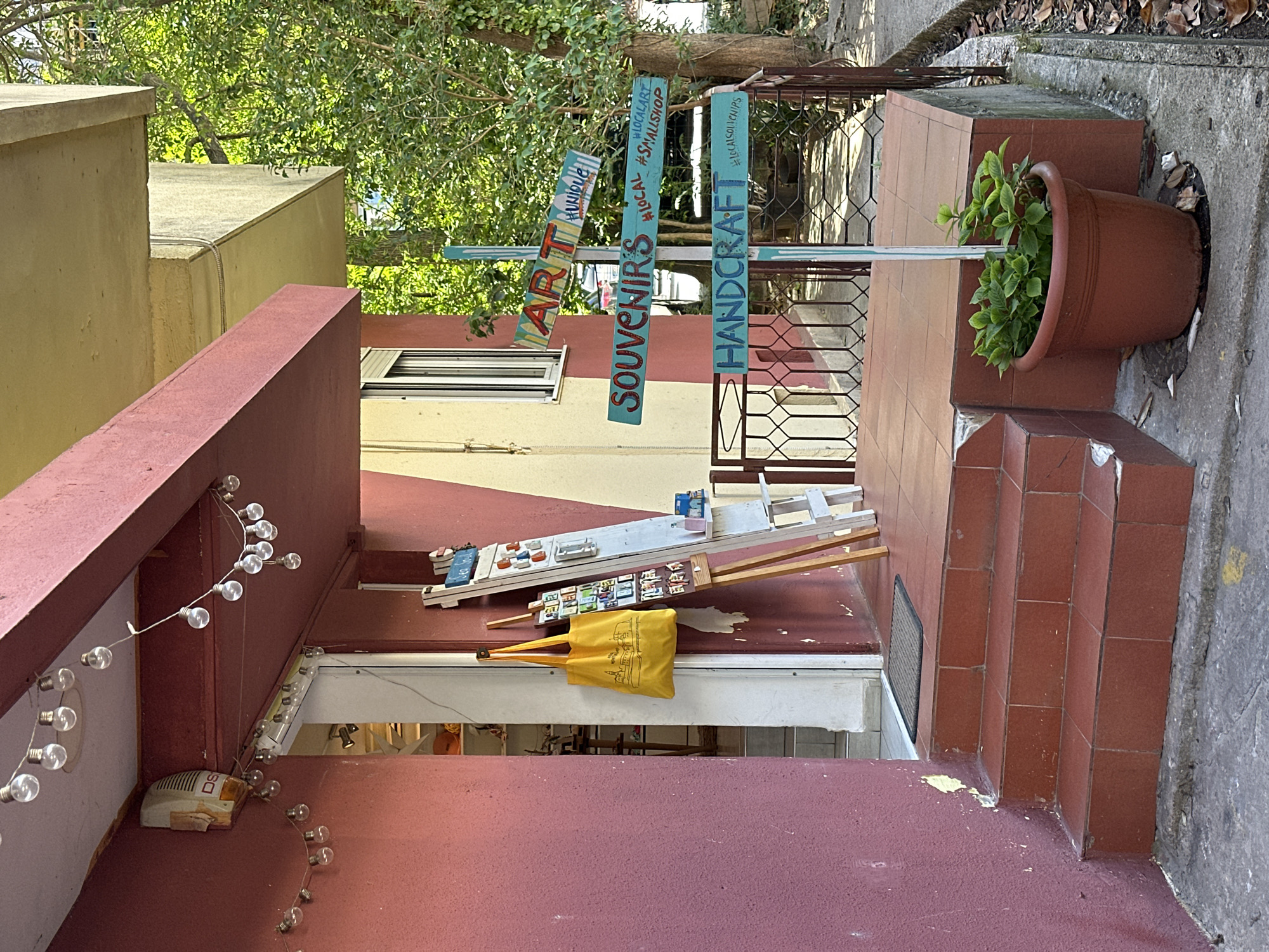 The image shows the entrance of a small shop in what is likely a tourist area. The shop is identified by a signpost with the words "ART", "SOUVENIRS", and "HANDCRAFT". The signs are painted in different colors and have a rustic appearance. The building has a two-tone color scheme of maroon and yellow, with the entrance area painted maroon. A string of lights hangs along the side of the building. Merchandise is displayed on a shelf above the entrance. A yellow bag hangs near the shelf. A pot with a green plant sits on the stairs leading up to the entrance. The steps are tiled. The environment suggests a location in a European city, consistent with Budva, Montenegro. The scene is quiet, with no visible people or activity. The building appears to be in a narrow street or alley.