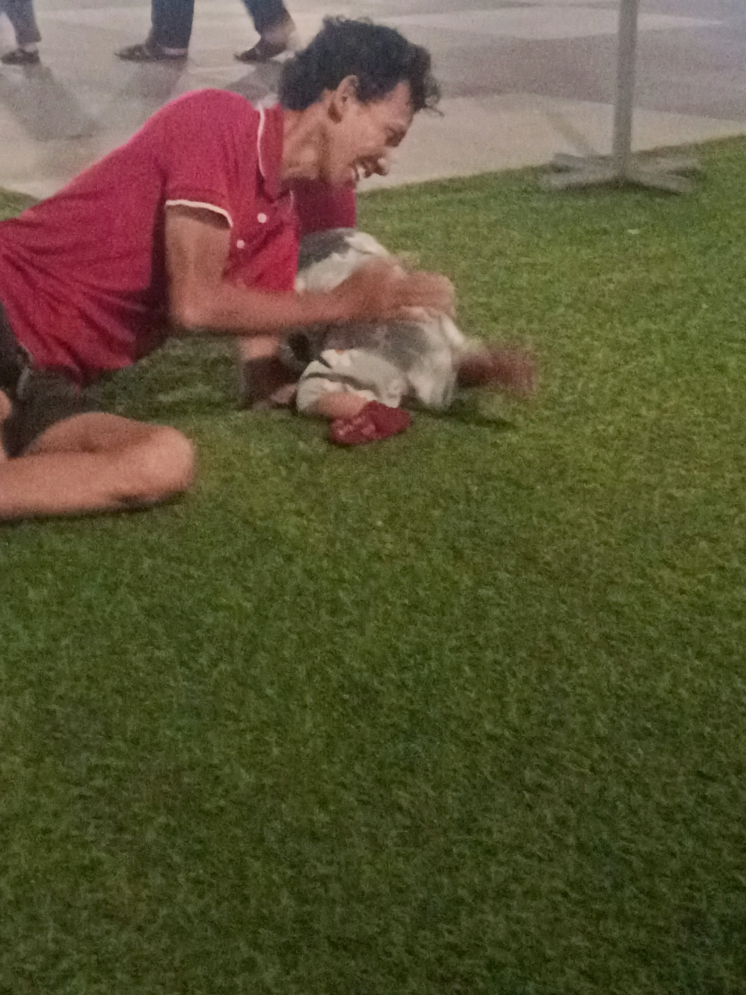 In a city park in Sidoarjo, Indonesia, a man wearing a red polo shirt and dark shorts is on the grass playing with a small child. The man is laughing and appears to be tickling the child. The child is wearing a striped shirt and red shoes. The background shows a paved area with a few people walking and a light pole. The ground is covered in artificial grass.