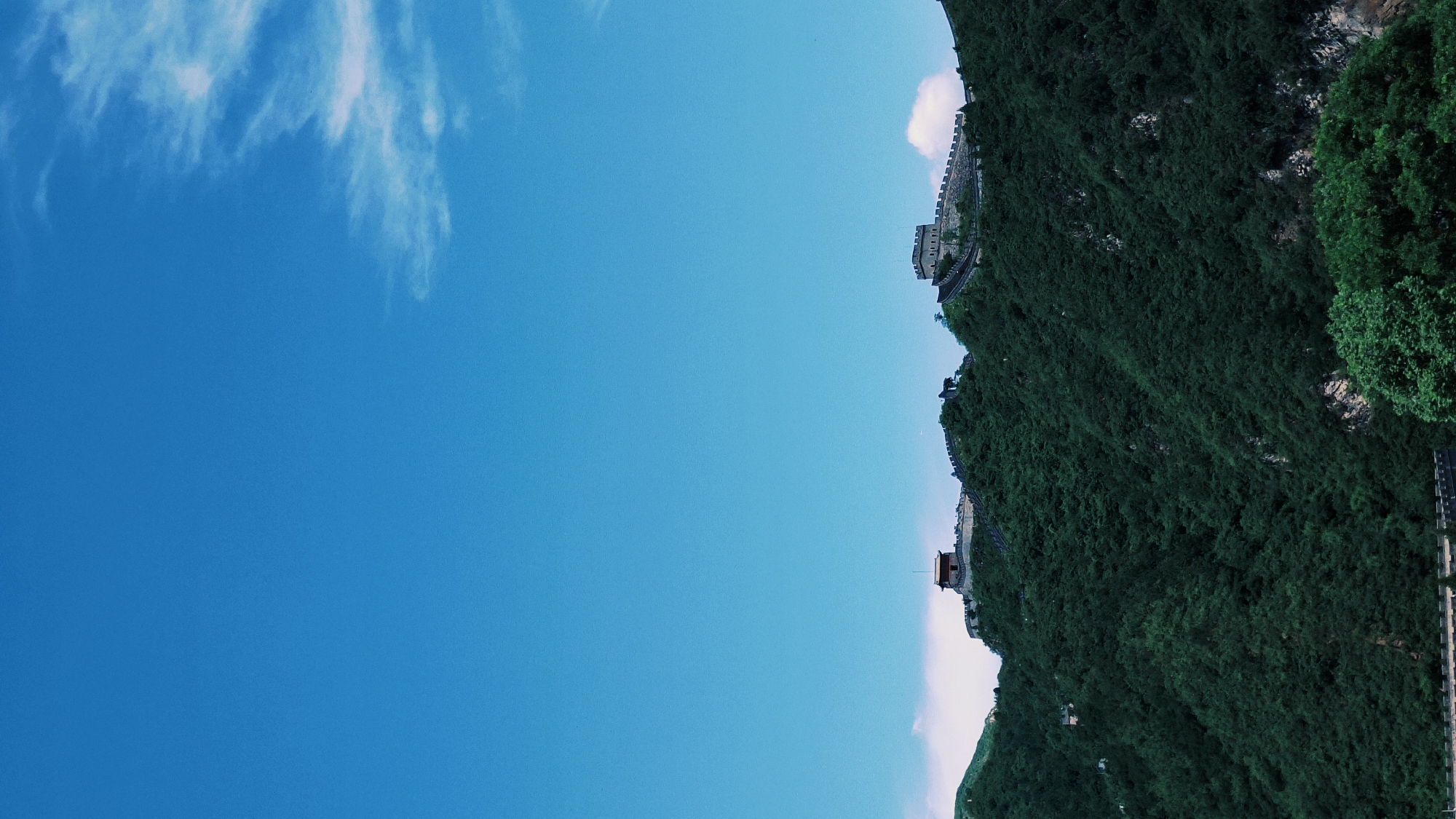 A wide-angle shot of a section of the Great Wall of China, positioned atop a steep, green, tree-covered mountainside under a blue sky with light white clouds in Changping District, China. The wall, constructed of light-colored stone, has several towers and battlements. The green mountainside is covered in dense vegetation.