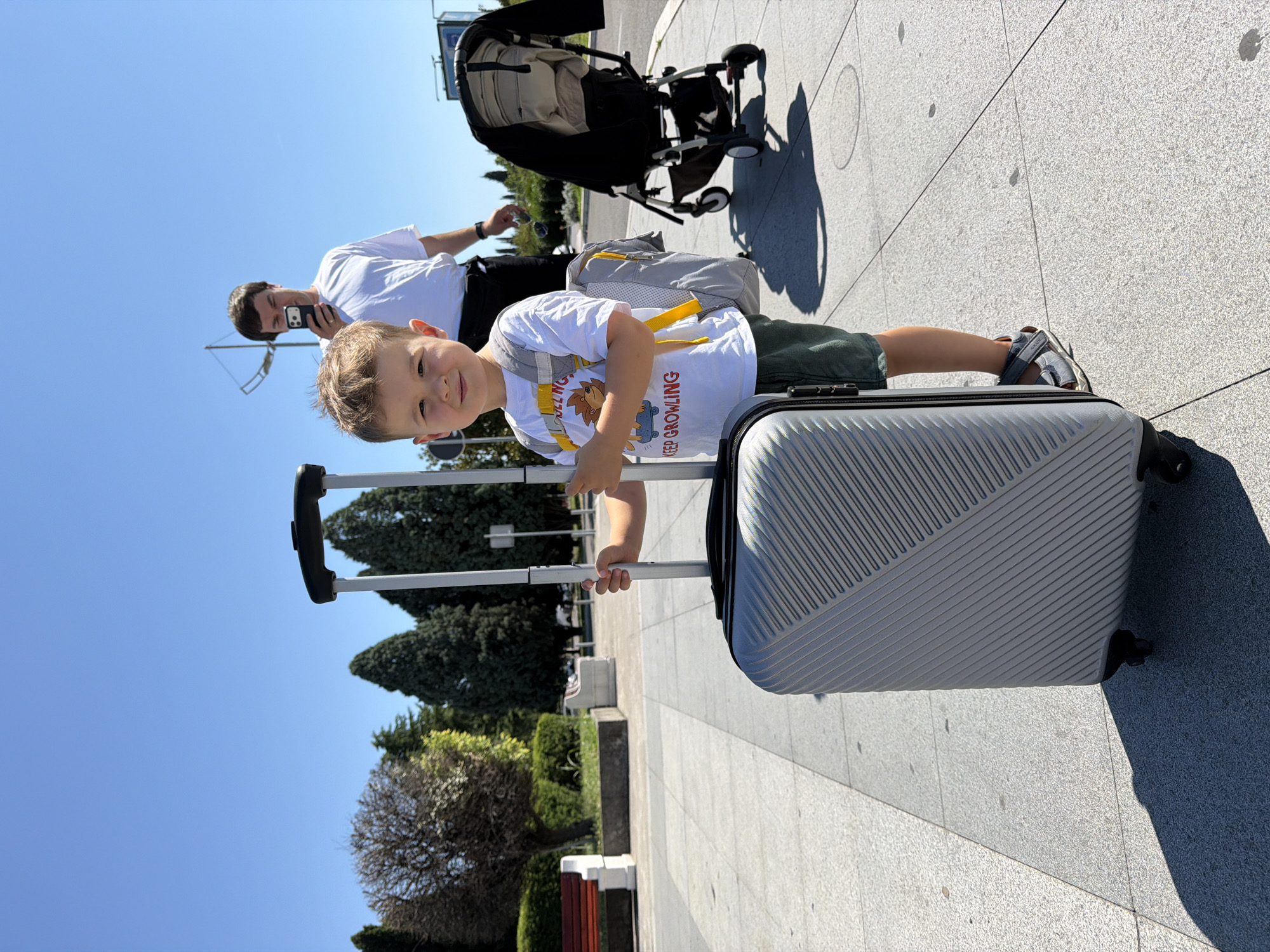 A young boy with blond hair and a white t-shirt with graphics and text stands on a paved area, holding the handle of a silver suitcase. He is wearing green shorts and sandals. Behind him, a man in a white t-shirt is looking at his phone, and a stroller is visible to the right. The background shows trees and benches, suggesting an outdoor public space. The scene appears to be in Donja Cijevna, Montenegro, under a clear blue sky.