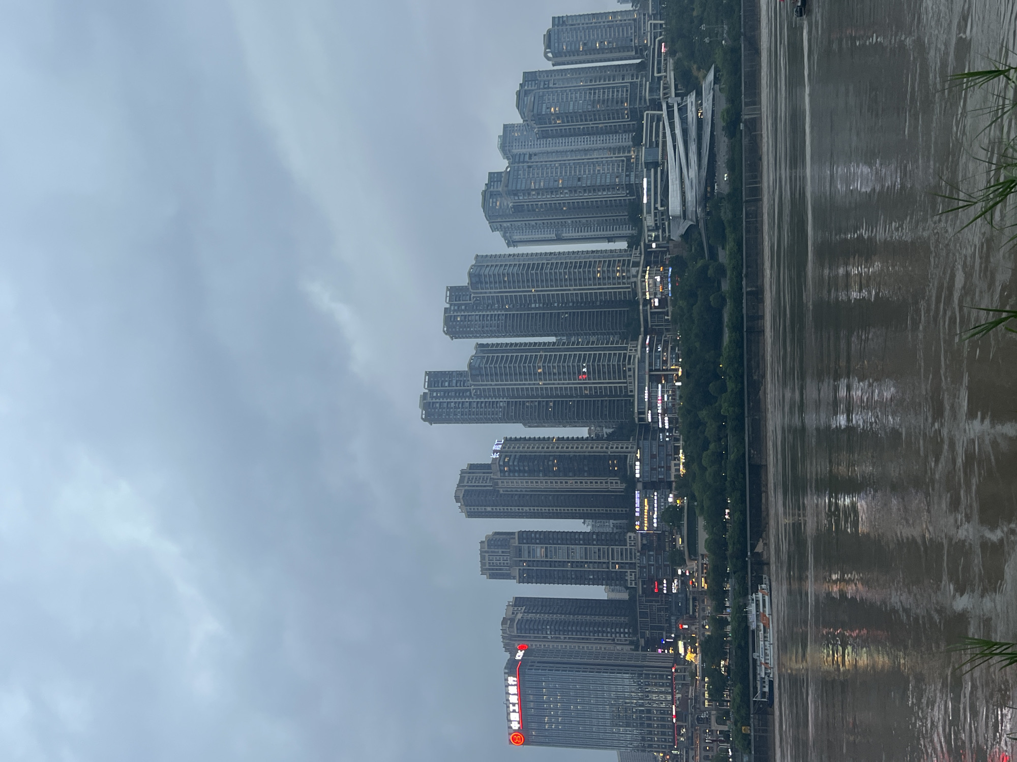 The image depicts a cityscape of Jiangbeicheng Street, China, dominated by a dense collection of high-rise buildings on a hillside overlooking a wide river. The sky is overcast and appears to be dusk or a cloudy day. The buildings are modern, with many glass facades, and some are illuminated by internal lights or external signage. The architecture varies, with some structures being slender and tall, while others are wider and more layered. At the base of the buildings, a complex of modern, angular structures is visible, possibly a cultural or commercial center. A wide, muddy river flows in the foreground, reflecting the muted colors of the sky and the lights from the city. A white and orange passenger boat is navigating the river, and its reflection is visible on the water's surface. Green vegetation is sparsely present along the riverbank and on the slopes behind the buildings. The overall impression is one of a bustling urban environment situated within a natural river valley.