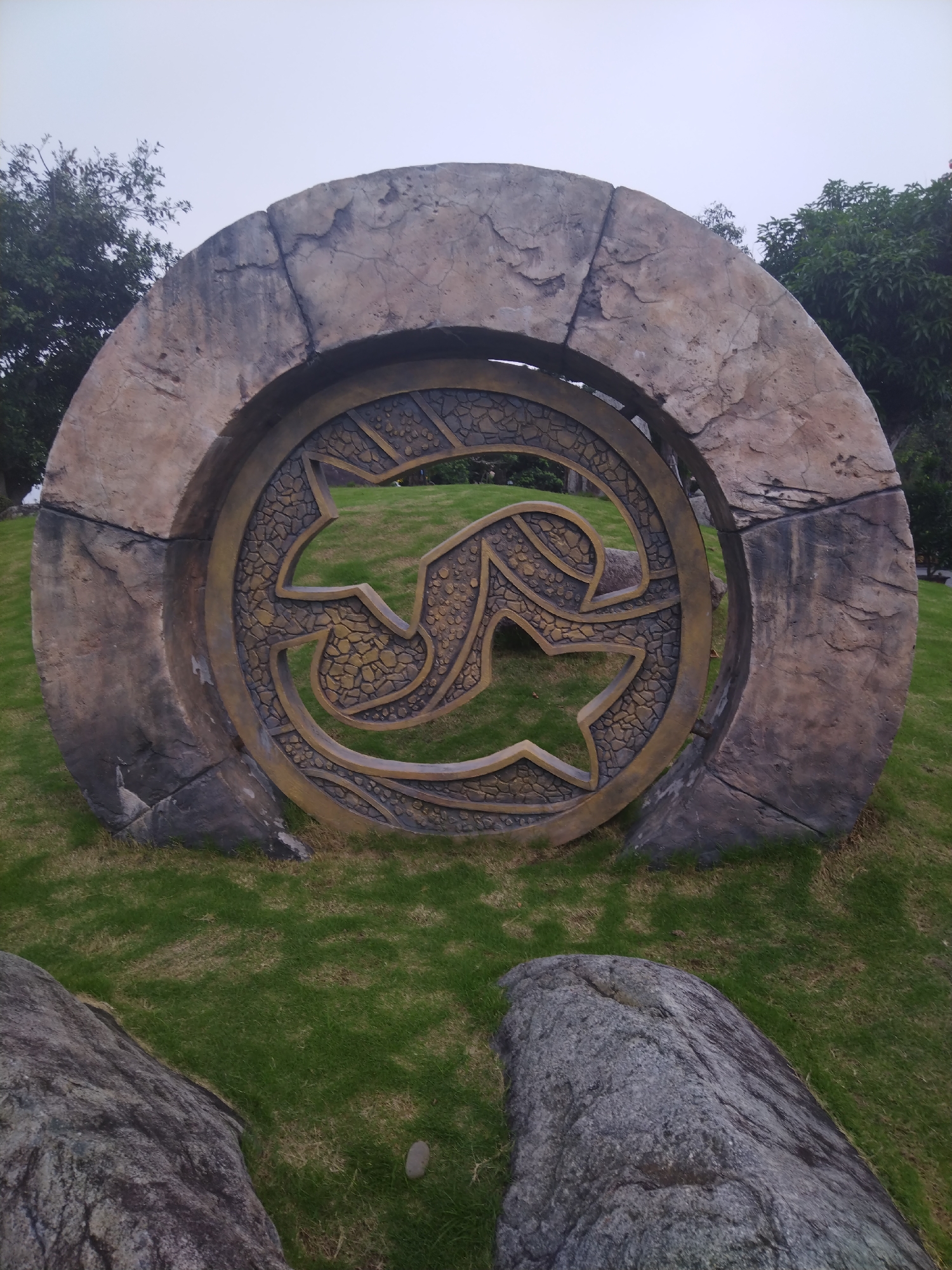 The image depicts a circular stone structure with an intricate design in the center, located outdoors in Hong Gai Ward, Vietnam. The structure appears to be a decorative or artistic installation, possibly situated in a park or garden setting. The surrounding environment includes a grassy area, trees, and other natural elements. Two gray rocks are visible in the foreground. The sky is overcast, suggesting cloudy weather.