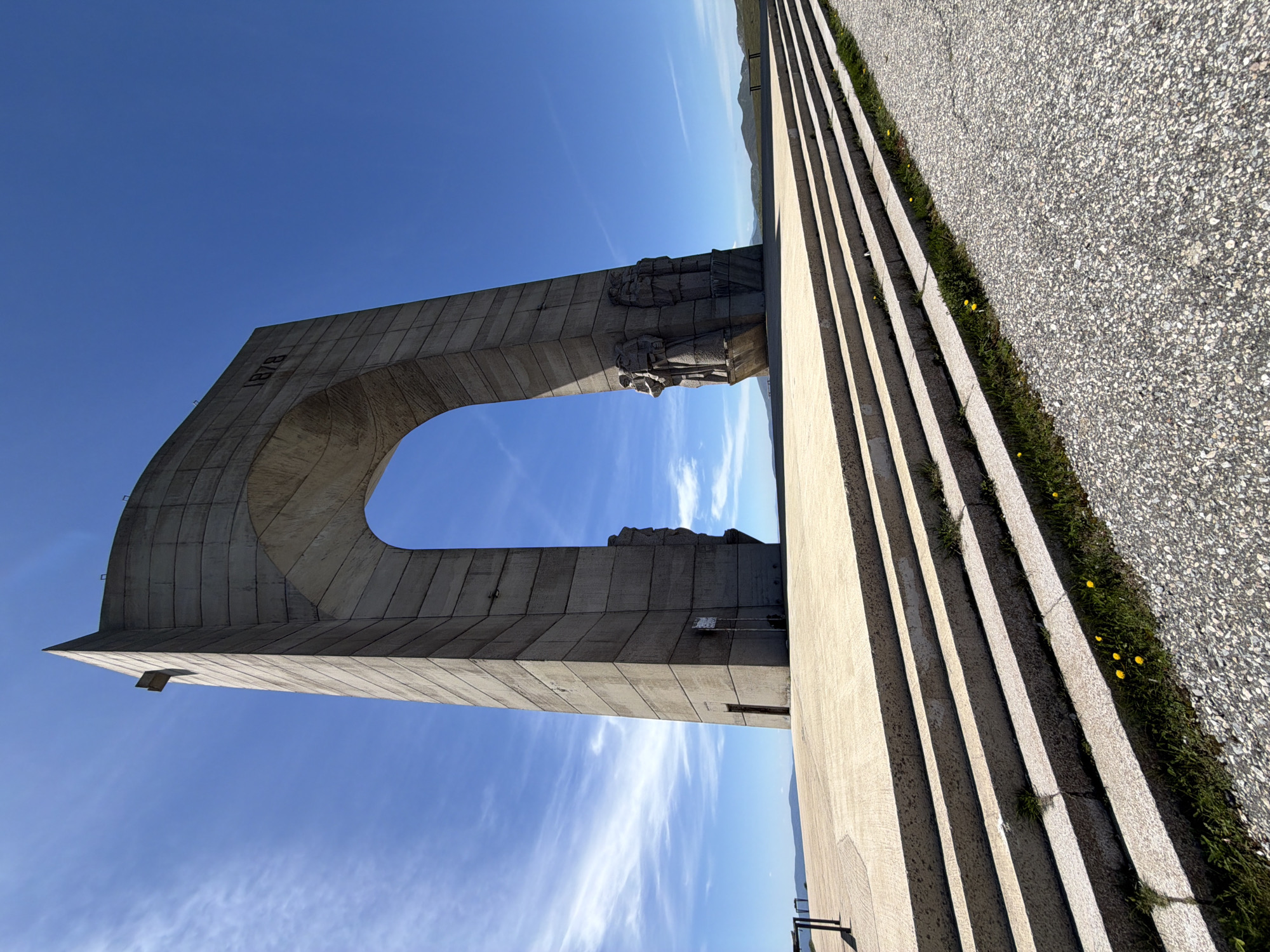 The image shows the Arch of Freedom Monument near Beli Osam, Bulgaria. The monument is constructed from gray concrete and features a large archway. The year "1878" is inscribed on one side. The arch also has some sculptures along the upper part. Beside it are large steps, and a stone path. The sky is blue with sparse clouds. The monument is set against a backdrop of mountains.