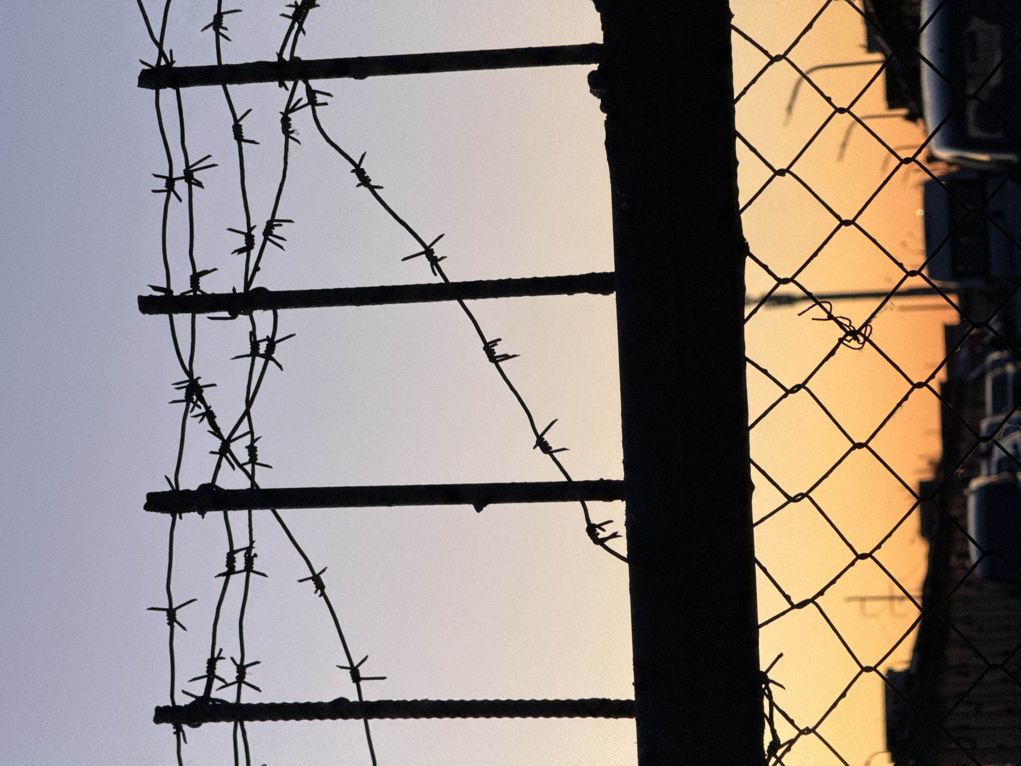 The image shows a close-up view of a fence with barbed wire. The fence is dark and silhouetted against a light, gradient sky, transitioning from a pale blue to a warm yellow-orange, suggestive of either sunrise or sunset. The barbed wire strands are intricate and detailed. To the right of the barbed wire is a more conventional chain-link fence. Behind this fence, blurred shapes suggest buildings. The photograph appears to be taken outdoors and is likely from Minsk, Belarus.
