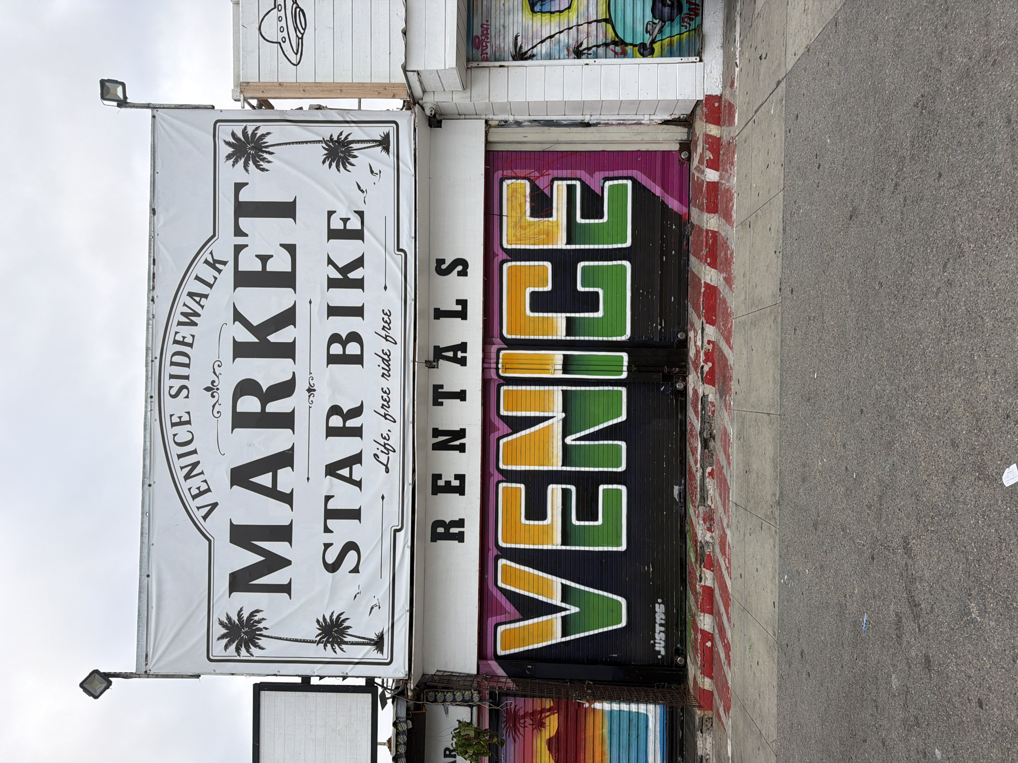 The image depicts the facade of a business on a street, likely in the Venice Beach area of Los Angeles, California, United States of America. The most prominent feature is a large white banner sign overhead with black text and graphics. The banner reads "VENICE SIDEWALK MARKET STAR BIKE Life, free ride free". Below the banner, a storefront features a roll-up metal gate painted with graffiti. The graffiti on the gate spells out "VENICE" in large, bold letters with a multi-colored fill of yellow, green, and purple, outlined in white. To the left and right of this main storefront, other businesses with graffiti-covered roll-up gates are visible, suggesting a vibrant commercial and artistic street scene. The ground in front of the storefront is a sidewalk with red and white striped paint on the curb and steps, indicating a designated crosswalk or traffic safety marking. The street itself is asphalt, with some debris scattered on its surface. The sky is overcast, suggesting a cloudy day. The overall impression is of a commercial area with a strong visual identity, characteristic of coastal tourist destinations.