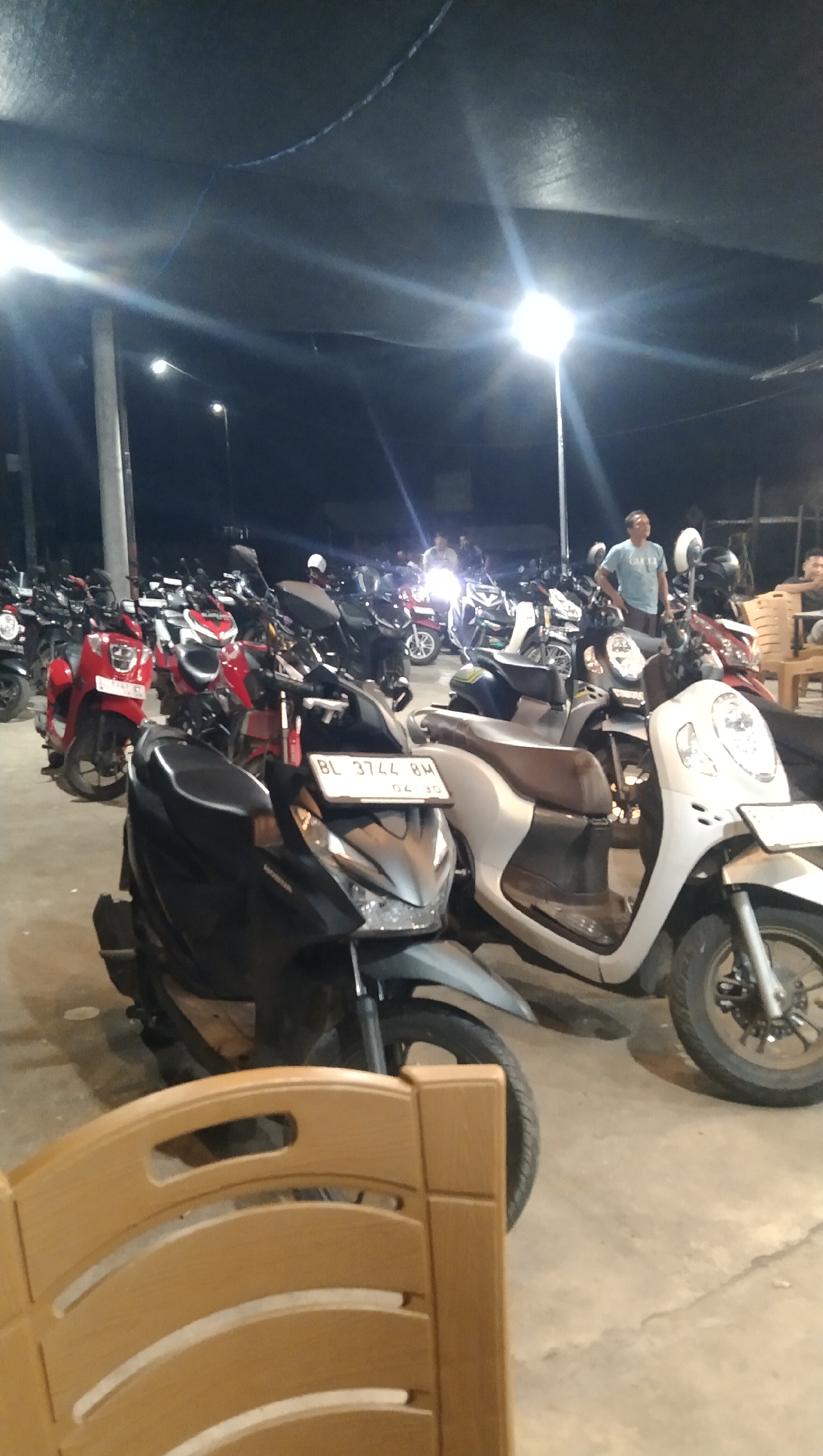 This media file captures a nighttime scene at a Keude Trienggadeng, Indonesia location, likely a parking area for motorcycles under a dark canopy. Several motorcycles are parked, predominantly scooters, in various colors including red, black, and white. The lighting appears to be from overhead lamps, creating bright spots and some glare.
In the background, a few people are visible, some standing and others possibly seated. One person, wearing a blue shirt and light-colored pants, is standing and looking towards the right of the frame. Another individual is partially visible in the lower right corner, seated on a wooden chair. The presence of parked motorcycles and people suggests a gathering or a place where people have congregated, possibly for an event or simply a communal space.
One of the motorcycles in the foreground has a license plate clearly visible with the text "BL 3744 BM" and possibly "04.30" below it. The ground is paved, appearing to be concrete. The overall atmosphere seems relaxed, with the lighting indicating it is nighttime. The scene is set outdoors, under a man-made shelter.