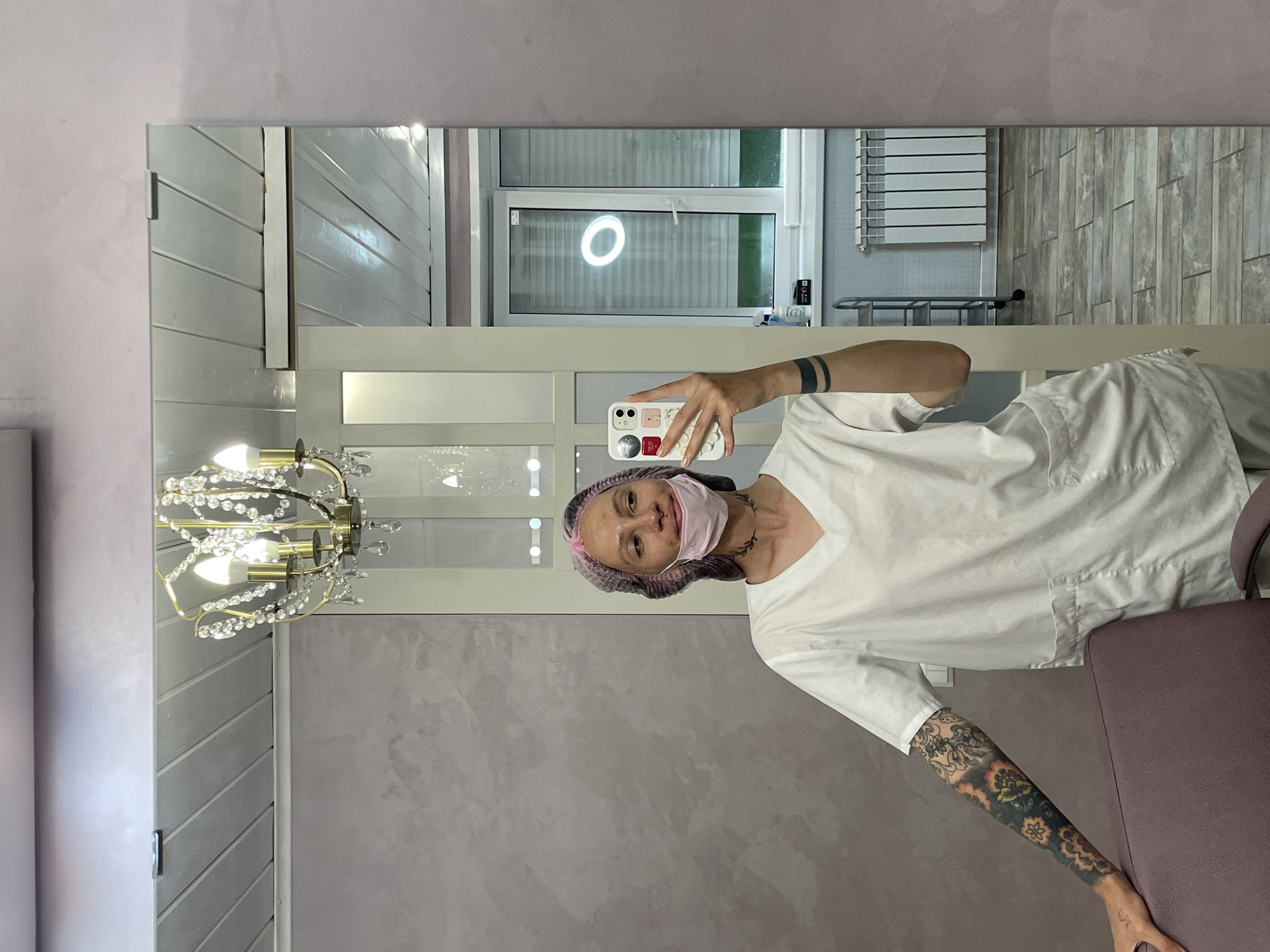 A person in a white medical uniform, a pink surgical cap, and a light pink face mask is taking a selfie in a mirror. The phone case is white with various colorful cartoon-like elements. The person has tattoos on their visible arm and around their wrist. The mirror reflects a room with light gray walls, a window with a round light fixture, a towel rack, and a chandelier. Based on the image, it appears to be an indoor setting, likely a clinic or salon. There is a chair to the right, near the bottom of the frame. The location is inferred to be Izhevsk, Russia, based on the given information.