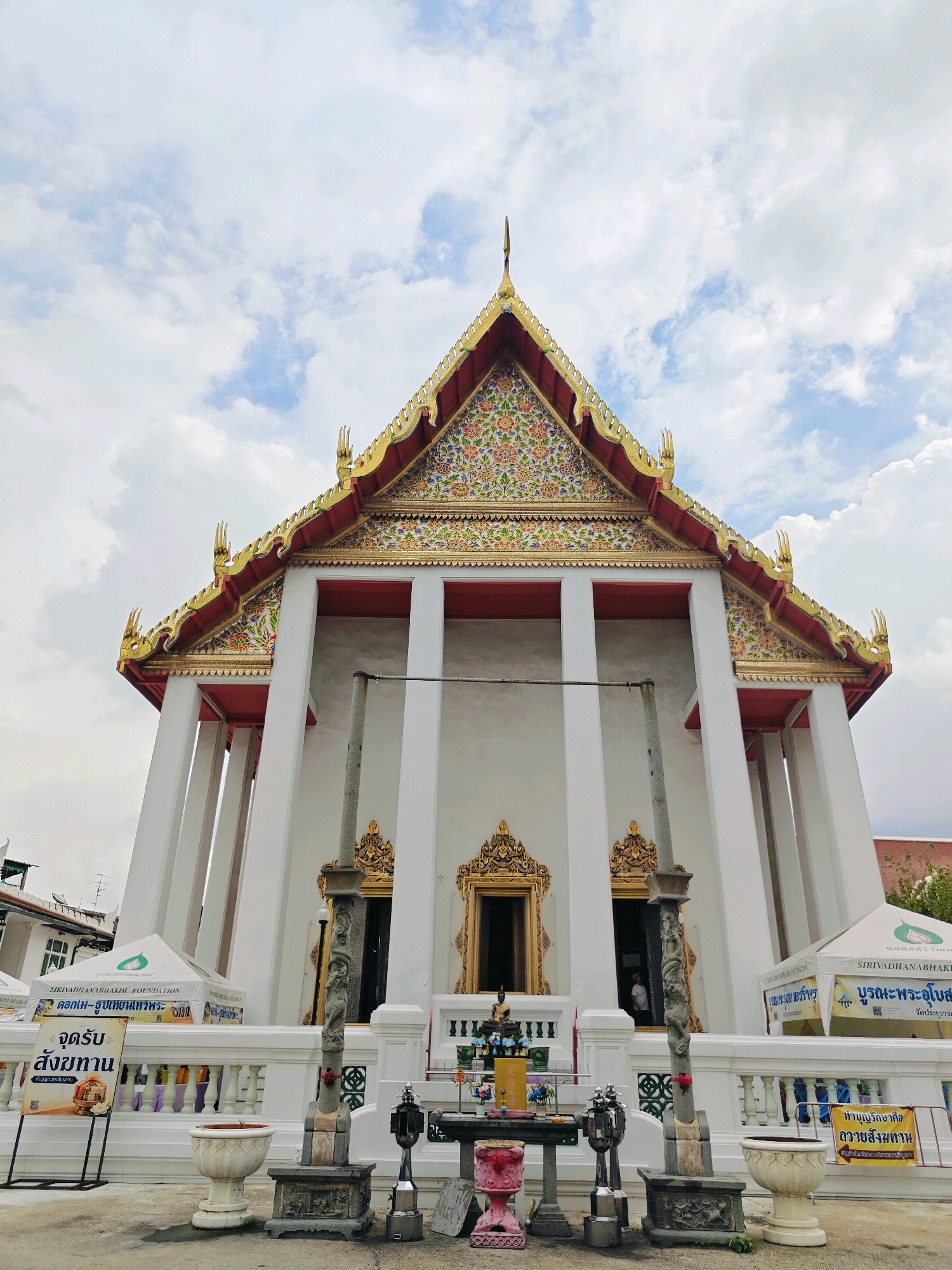 The image captures the exterior of a white temple building with a prominent, ornate golden roof adorned with intricate floral patterns. The temple has large white columns supporting the roof, and ornate golden decorations flank the entrances. In front of the temple, there is a white railing. To the left and right of the main structure are white tents with logos and Thai text. In the foreground, there is a paved area with several objects including decorative planters, a stone pillar with carved figures, a table with offerings such as flowers and incense, and various metallic objects. A yellow sign with Thai text is placed on the left side of the paved area. A person can be seen partially visible through one of the temple doorways. The sky above is cloudy. Based on the architecture and the presence of Thai script, the image is likely taken at a temple in Bangkok, Thailand.