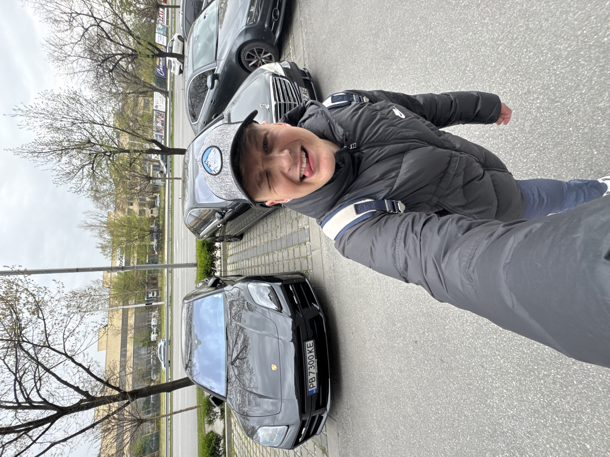 The image is a selfie, with the photographer tilting the camera. A person with a cap and a gray jacket is in the foreground, sticking out their tongue. Behind them are parked vehicles, including a black Porsche Urus with the license plate "PB 7300 KE". The background includes trees and a building, suggesting an urban area. Based on the license plate and context clues, the location is likely Plovdiv, Bulgaria.