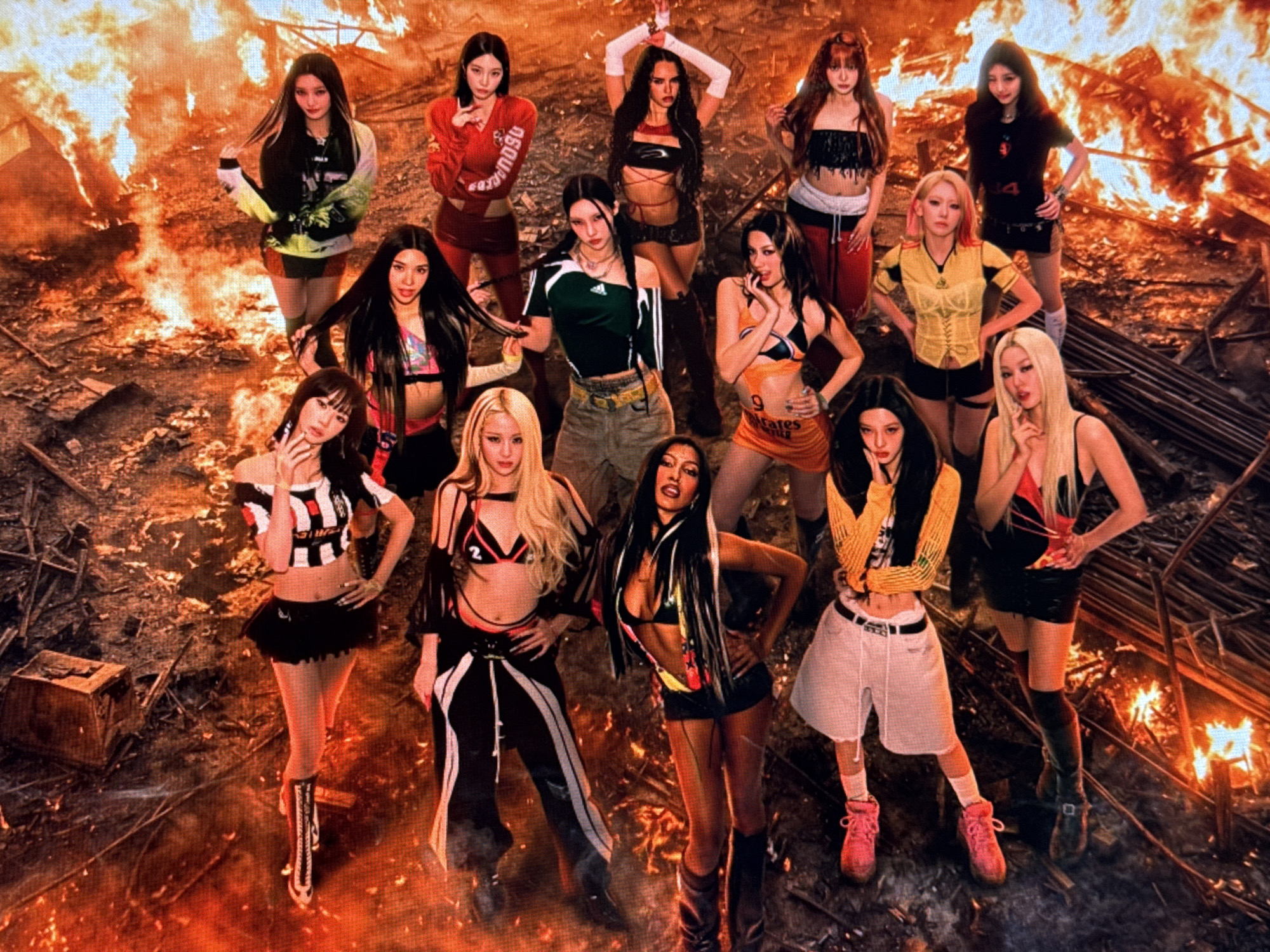 The image depicts a group of thirteen young women posing in a gritty, post-apocalyptic urban environment with visible flames and debris. The women are diverse in appearance and fashion, with some wearing dark, edgy outfits, while others are in more colorful and sporty attire. Their poses suggest a deliberate and confident presentation. The background features intense fire, scattered wooden structures, and what appears to be rubble, creating a dramatic and hazardous atmosphere. The overall scene is dimly lit, with the fire providing the primary source of illumination, casting warm, orange hues across the setting and the subjects. There is no discernible presence of the city of Baku, Azerbaijan in this image; the scene is entirely focused on the group and their immediate, fiery surroundings.