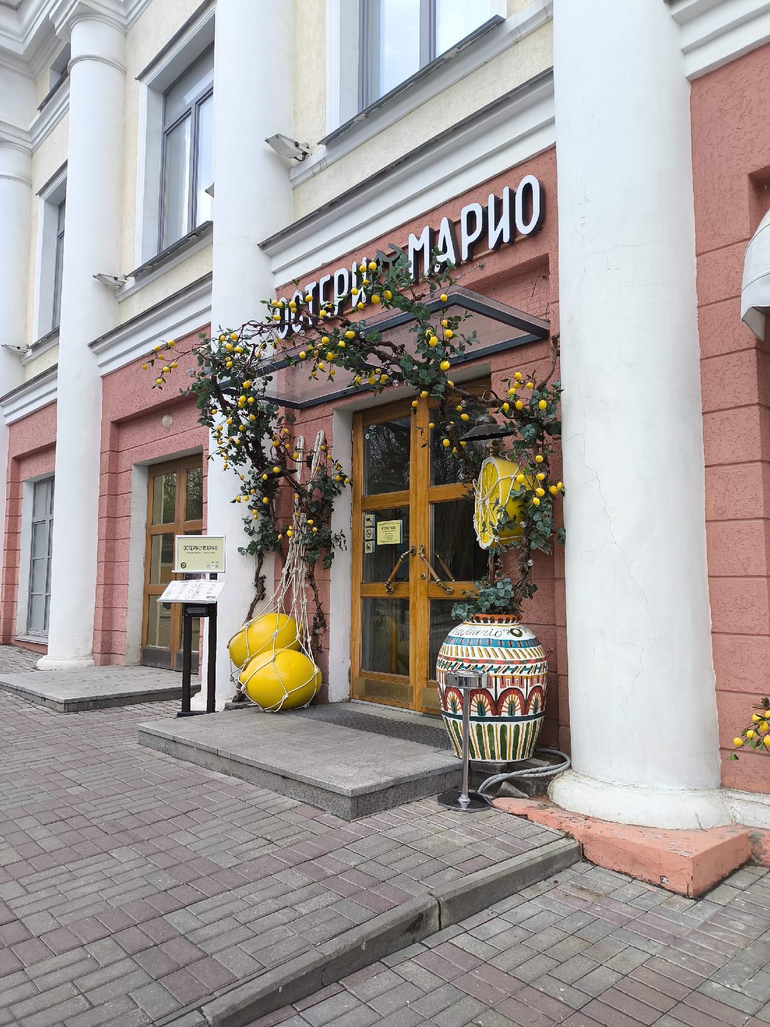 The image shows the entrance to a business establishment called "OSTERIA MARIO" in an urban setting. The building's facade is adorned with white columns and pink walls. A decorative lemon tree with artificial lemons is wrapped around the entrance, and a pair of oversized yellow lemon-shaped objects are suspended in a net next to the door. A colorful, patterned vase sits near the entrance, next to one of the columns. The entrance has a glass awning, and the doors are made of wood. The establishment is situated on a paved street with a herringbone brick pattern. The location appears to be a city, but the exact city and country are unknown.