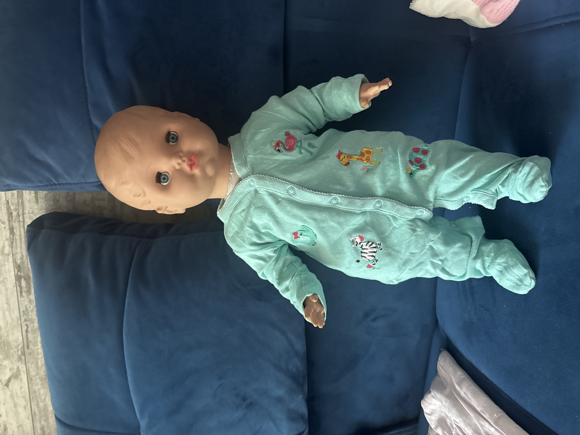 A life-sized baby doll is positioned upright on a dark blue, tufted sofa. The doll wears a light teal, footed onesie adorned with embroidered cartoon animals, including a flamingo, a giraffe, a zebra, and a turtle. The doll has light-colored hair, blue eyes, and pink lips. To the left of the doll, a folded piece of light-colored fabric, possibly clothing, rests on the sofa. To the right, a white and pink sock is partially visible on the sofa. The background consists of a textured, gray surface that appears to be a wall. The scene is illuminated by overhead lighting. There is no indication of the city of Kokshetau, Kazakhstan within the image.