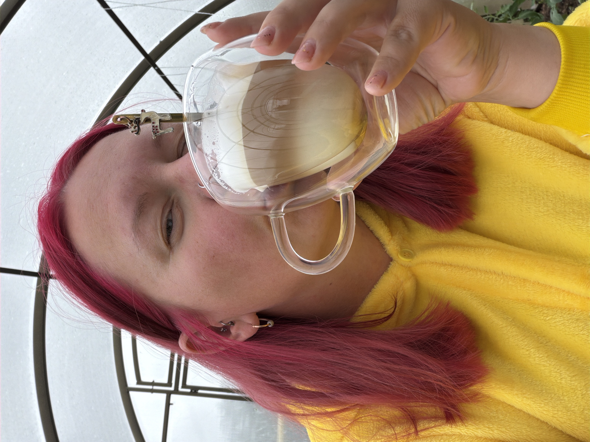 An individual with bright fuchsia-colored hair and dressed in a yellow plush garment is depicted indoors, in what appears to be a greenhouse or conservatory, attributed to the city of Chaikovsky, Russia. The person is actively drinking from a clear, double-walled glass mug, held in their right hand. The mug contains a layered beverage, consistent with a latte or cappuccino, featuring distinct creamy white, light brown, and foamed white layers. A small metallic spoon, topped with a decorative white reindeer figurine featuring red spots and brown antlers, is resting within the drink. The subject has fair skin, with their right eye (blue/grey iris) visible and directed towards the camera. A small gold-colored hoop earring is present in their right earlobe. Their fingernails are manicured with light pink polish and subtle glitter specks. The background shows transparent or translucent panels supported by dark, arched structural framing, through which an overcast sky is visible.