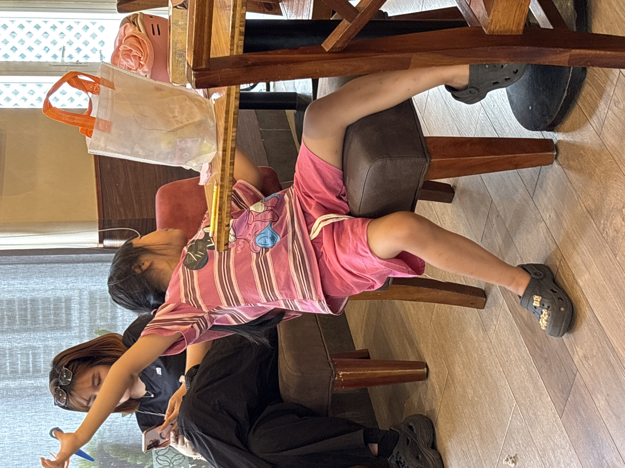 The image depicts an indoor scene, likely a cafe or restaurant, located in Nha Trang Ward, Vietnam. In the foreground, a young girl is seated on a dark brown upholstered chair, facing away from the camera. She is wearing a pink horizontally striped t-shirt with a cartoon character motif and pink shorts. Her right arm is extended upwards and to the left. She is wearing dark grey Croc-style shoes. To her left, a woman is seated on a dark brown upholstered chair, also facing away from the camera. She is wearing a black t-shirt and black pants. She is holding a pink smartphone in her left hand and appears to be looking at it. Her right arm is extended and holding a blue object, possibly a pen.
In the background, a wooden table is visible, with a white translucent shopping bag with orange handles placed on it. The bag contains various items. To the right of the bag is a pink molded plastic container with a lid, holding what appears to be folded fabric. Behind the table, a large window with a diamond-patterned glass is partially visible, suggesting natural light is entering the space. The floor is tiled with a wood-grain pattern. The overall setting appears casual and relaxed.