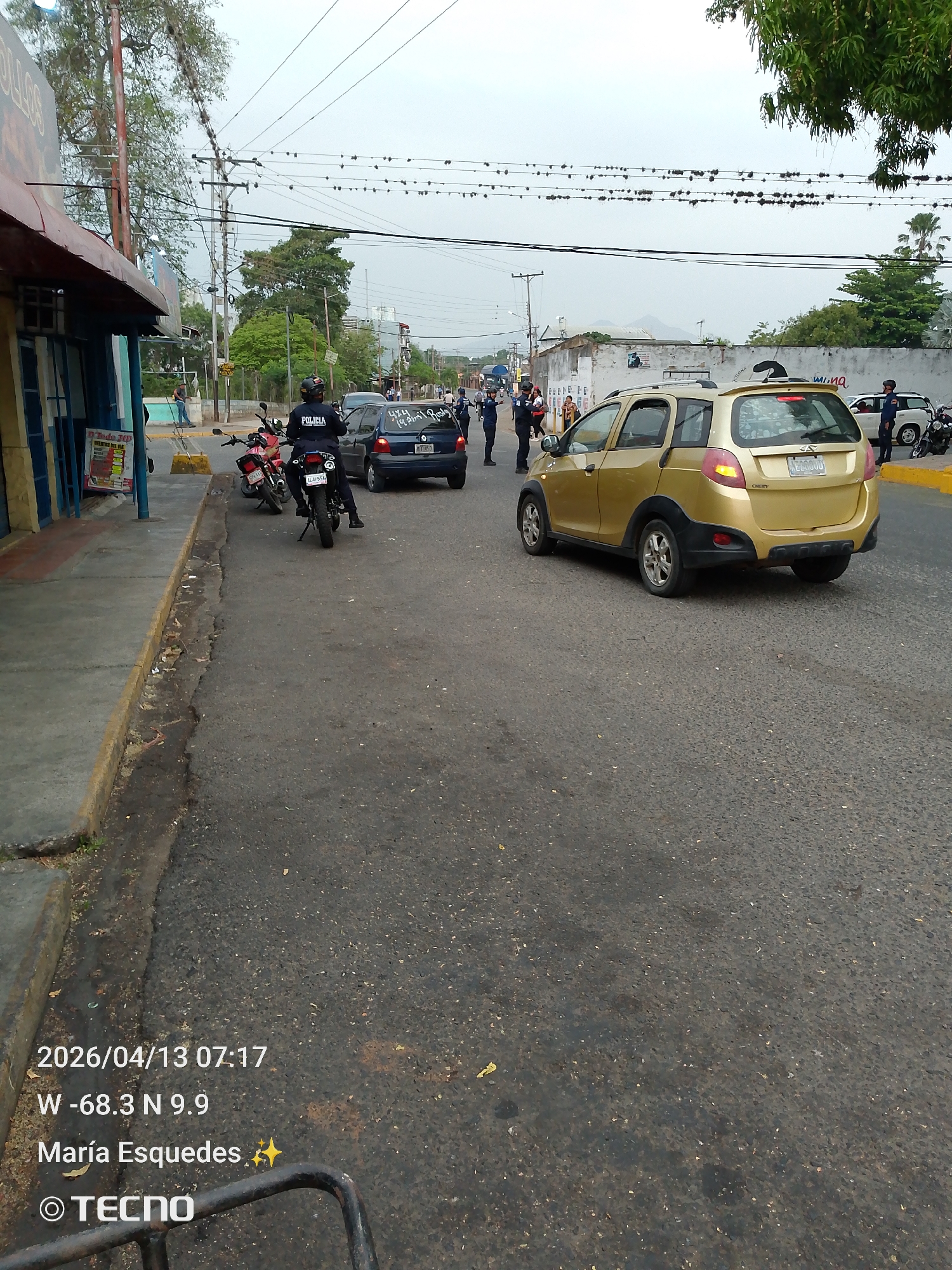 The image depicts an urban scene in Tinaquillo, Venezuela, taken on April 13, 2026, at 07:17. In the foreground, a gold Chery vehicle is positioned on the right side of a paved road. A police officer on a motorcycle is stopping a dark blue compact car in the distance. Additional uniformed officers are visible on the right side of the road, potentially interacting with other vehicles and people. Along the left edge, a building fronts the sidewalk. Power lines are above.