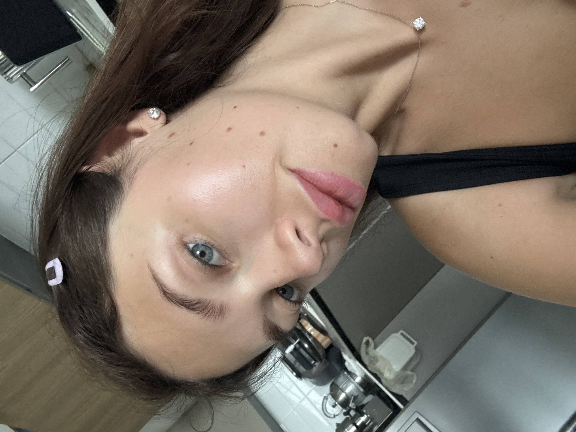 A woman with brown hair and blue eyes is visible. She has a small, light purple hair clip in her hair. She is wearing a diamond stud earring and a necklace with a diamond pendant. She is wearing a black strapless top. The background shows a kitchen area with white tile walls and stainless steel appliances. This location cannot be confirmed as Tel-Aviv, Israel, with the provided information.