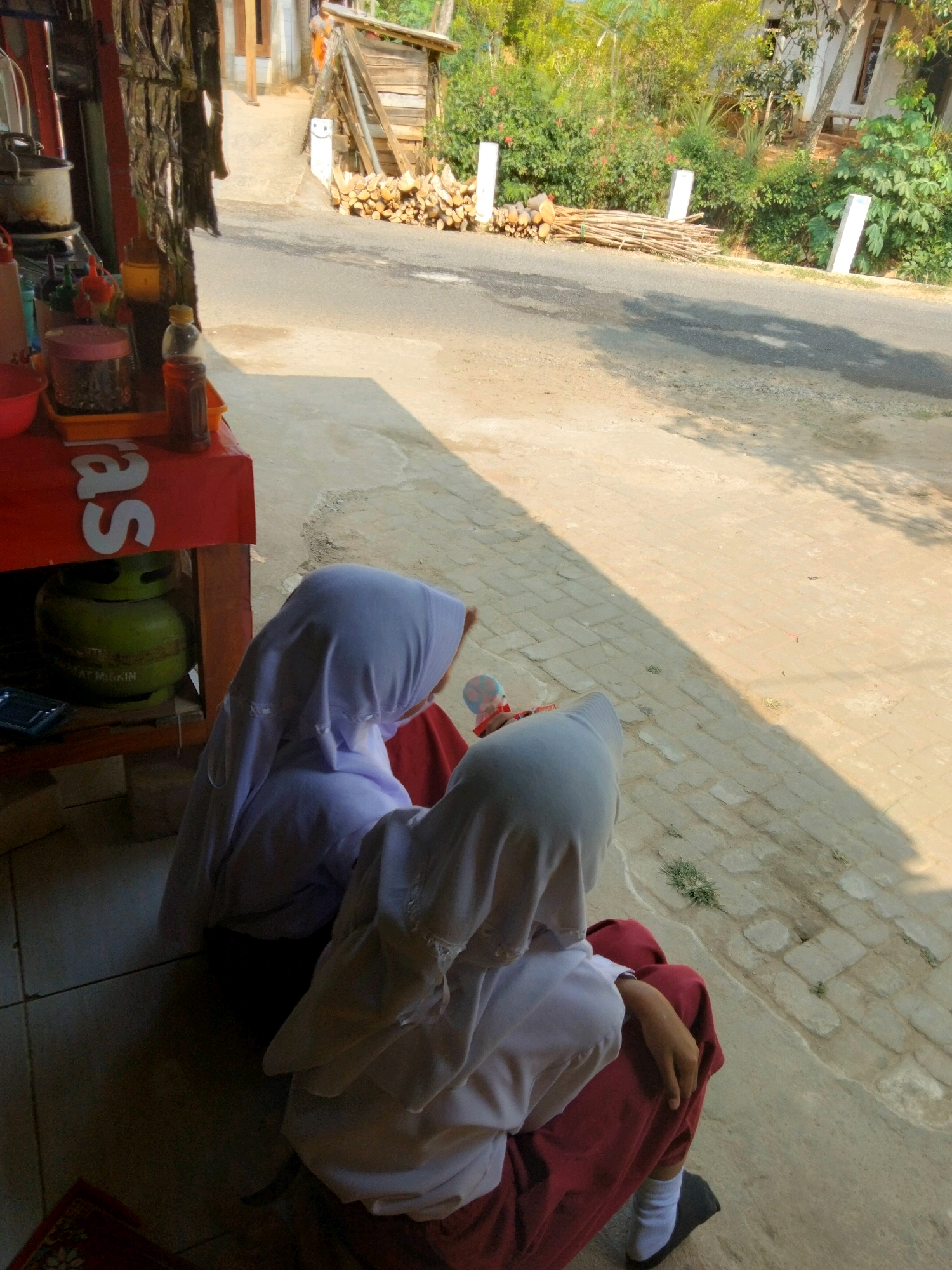 The image shows two young girls wearing hijabs and red skirts sitting on the ground in front of a food stall. The stall has a red counter with various items on display, including jars and bottles of condiments. A green gas cylinder is visible underneath the counter. The scene is set on a sunny day with a road and lush vegetation in the background. The location is identified as Sidorejo, Indonesia. The girls appear to be either customers or helpers at the stall.