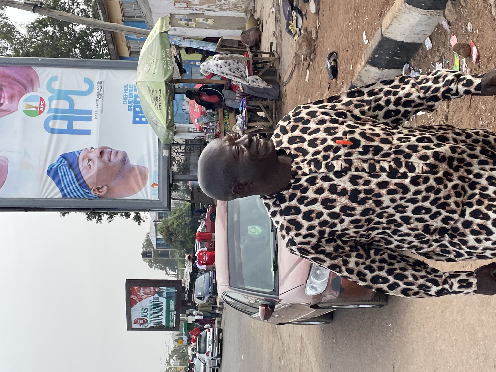 The image depicts a man in a leopard-print shirt standing near a street in Jos, Nigeria. A car is parked nearby. In the background, there are billboards promoting the APC party and a candidate named Caleb Manasseh Mutfwang for governor, as well as Chief Hon David Bitrus. An Outpouring billboard can be seen near the car. Other individuals and shops are also visible, alongside traffic on the road.