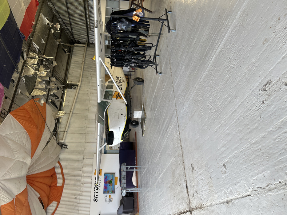The image displays the interior of a skydiving facility located in Chascomús, Argentina. The foreground consists of a light grey concrete floor exhibiting wear and visible seams. In the midground, a single-engine propeller aircraft, predominantly white with yellow accents and "SKYDIVE CENTER" branding on its fuselage, is parked. The aircraft's nose cowling is detached, exposing internal engine components. It is positioned on a grey block beneath its front landing gear and a metal plate under its rear wheel. To the left of the aircraft, a lounge area is arranged against a white wall, featuring a dark grey armchair, a dark blue couch, and a low, light-colored table. A television screen on the wall displays an animated cartoon image, and a "SKYDIVE CENTER" logo with a QR code is also visible. To the right of the aircraft, a mobile, black metal rack holds multiple black skydiving harness rigs. The background reveals corrugated metal walls and a high ceiling structure. Numerous large, colorful parachutes (including orange, white, blue, green, yellow, and red) are partially unfurled or draped from the ceiling, alongside decorative or insulating foil-like materials and exposed industrial light fixtures. No individuals are present within the scene.