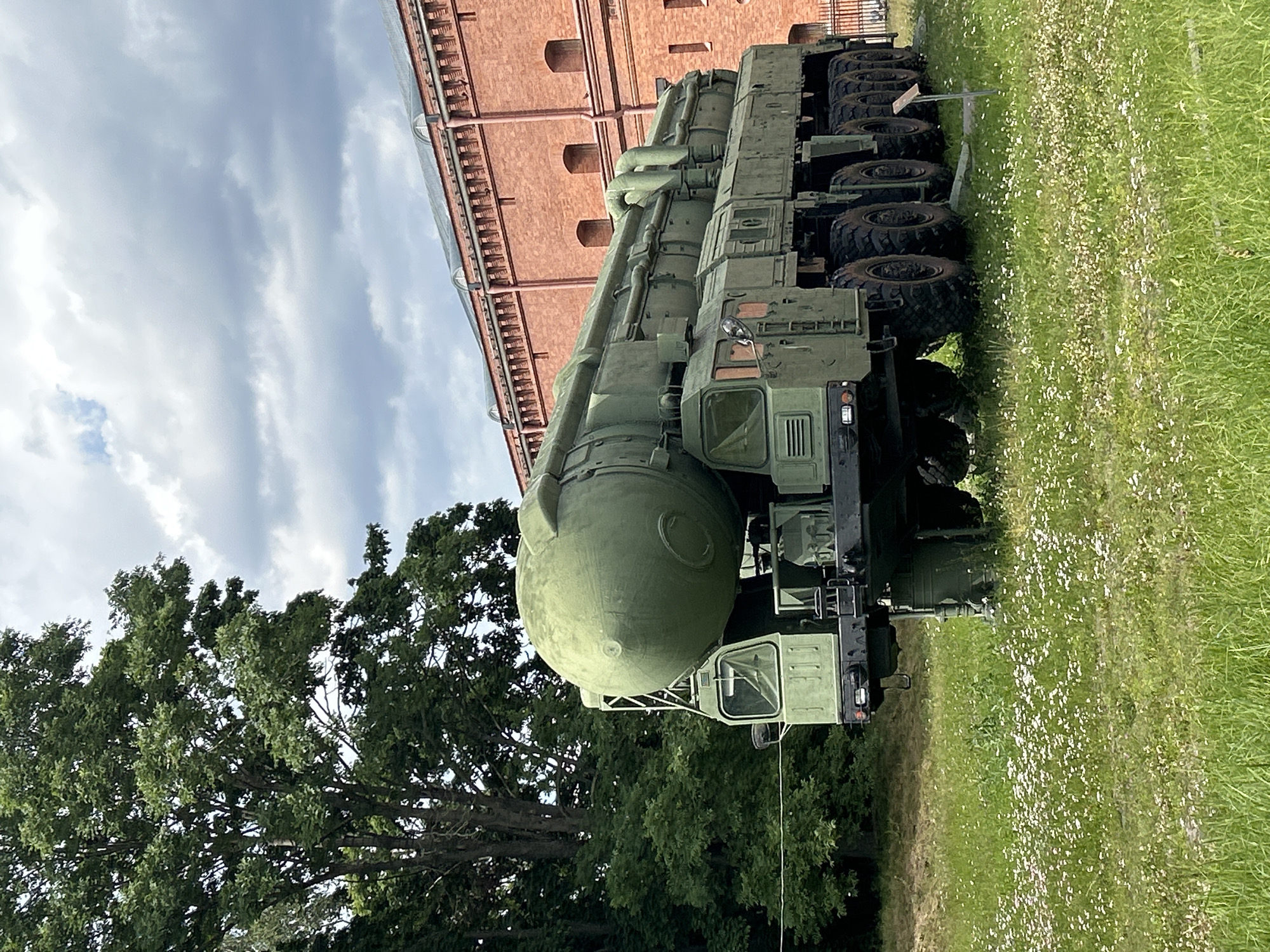 An outdoor scene shows a large, green, military missile launcher parked on a grassy area with scattered white wildflowers. The missile launcher is a multi-wheeled vehicle with a large missile situated on top. Behind the vehicle, a large, red brick building with arched windows and a dark roof is visible. To the left of the vehicle, a large tree with dark green foliage stands. The sky is cloudy with patches of blue.