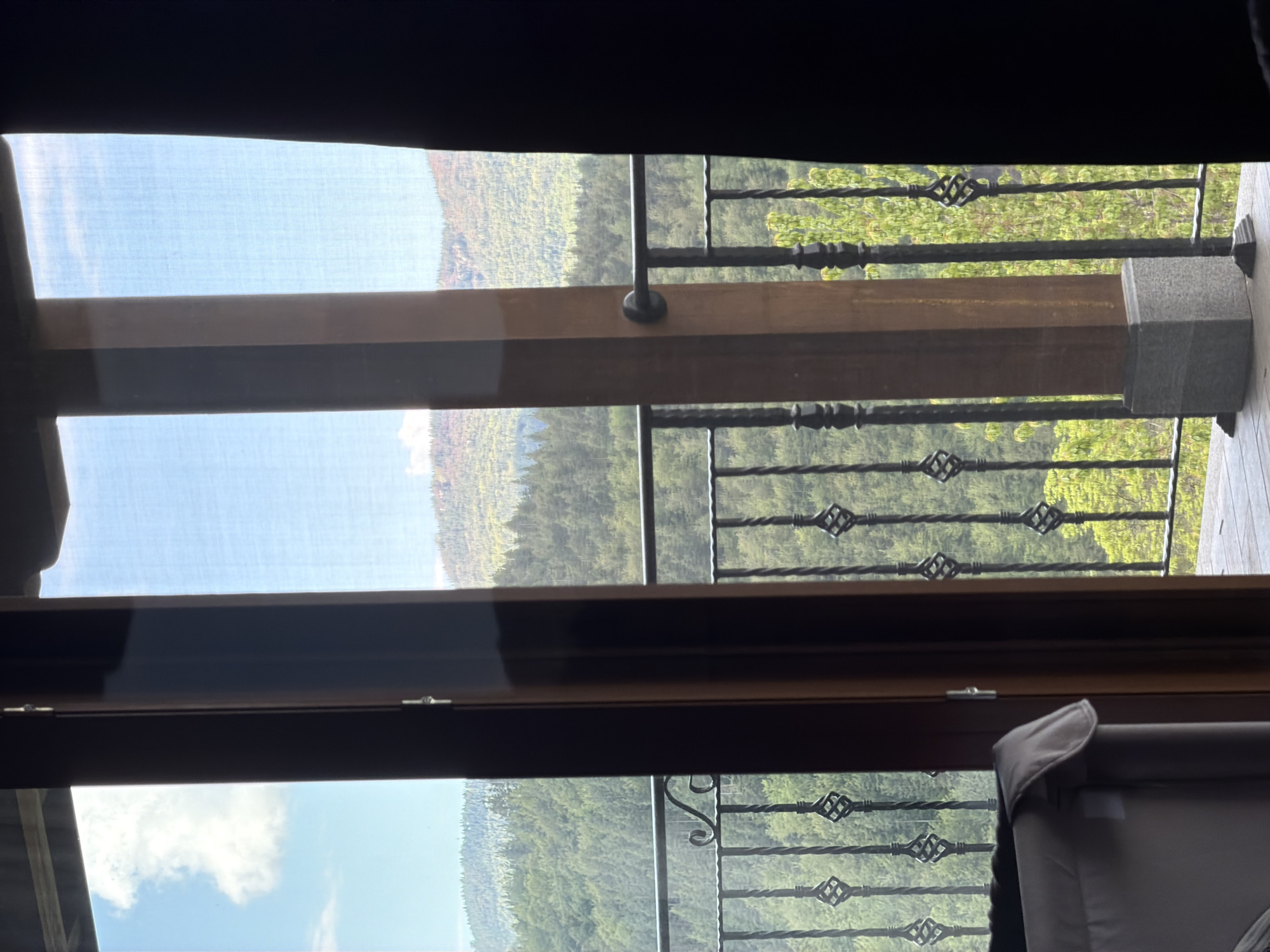The image presents a view from inside a building, looking out through a window with a balcony. The balcony has a decorative wrought-iron railing. The foreground includes elements of the window frame and a partially visible roller blind. Outside, there is a forested area, likely a mountainous or hilly region, as it has a hilly topography. The presence of trees suggests a natural environment, and there are no visible indications of urban development. The light conditions appear to be either daytime or early evening, suggesting that there is likely sunlight. The location given, Кирково, Bulgaria, is in a mountainous region, and the photograph is consistent with a setting that is set in or near a natural setting.