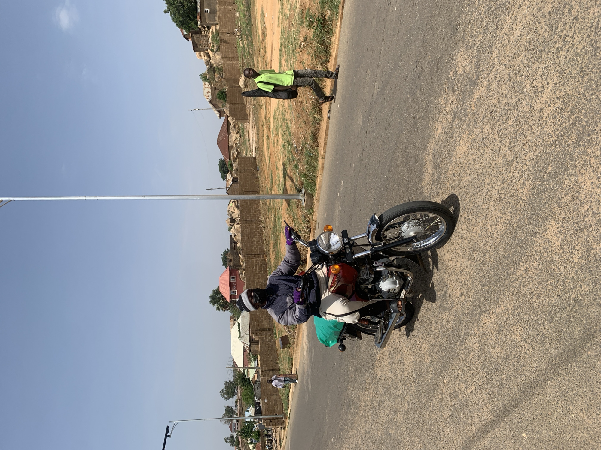 A man on a motorcycle is stopped on an asphalt road in Gura Top, Nigeria. He is wearing a dark jacket, light-colored pants, gloves, and a helmet. A green bag is slung over the motorcycle. Behind him, another man in a green shirt and dark pants walks along the side of the road. The background includes buildings and vegetation typical of the region. The sky is clear. Utility poles are visible along the side of the road.