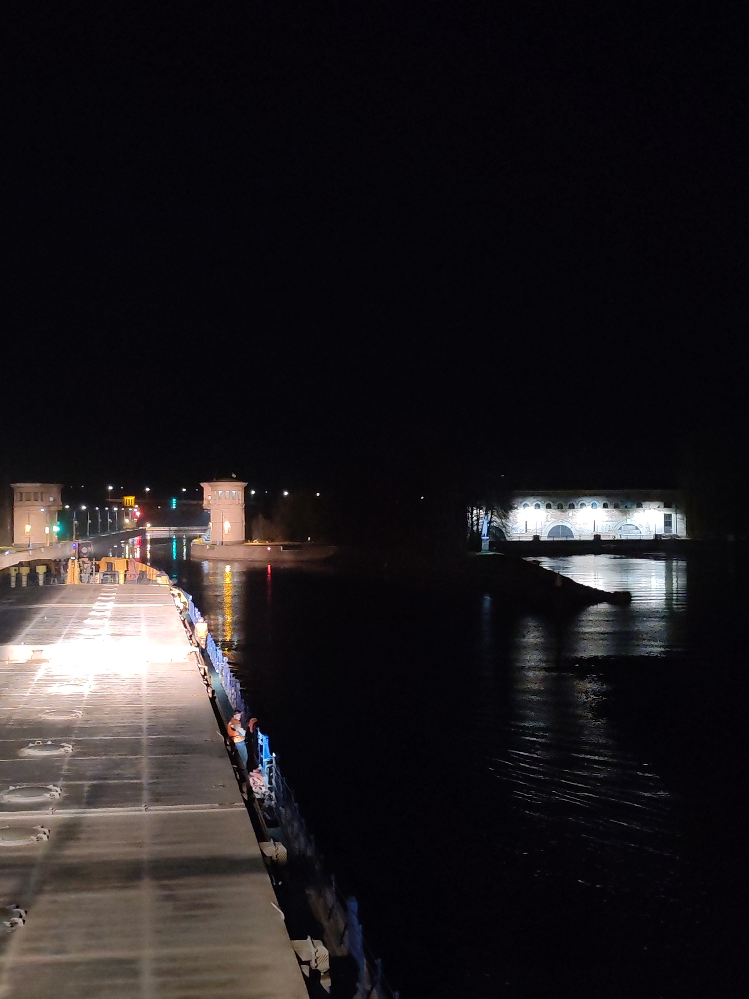 The image depicts a night scene in Базарово, Russia, showing a waterway with structures on either side. On the left, two small towers flank what appears to be a canal lock or bridge illuminated by streetlights. Reflections of these lights are visible in the water. To the right, a brightly lit, arched building sits at the edge of the water. The image is taken from the deck of a ship or platform with deck features visible in the foreground. People are present on the ship, standing by the railings. The overall environment is dark, with artificial light sources illuminating specific areas.