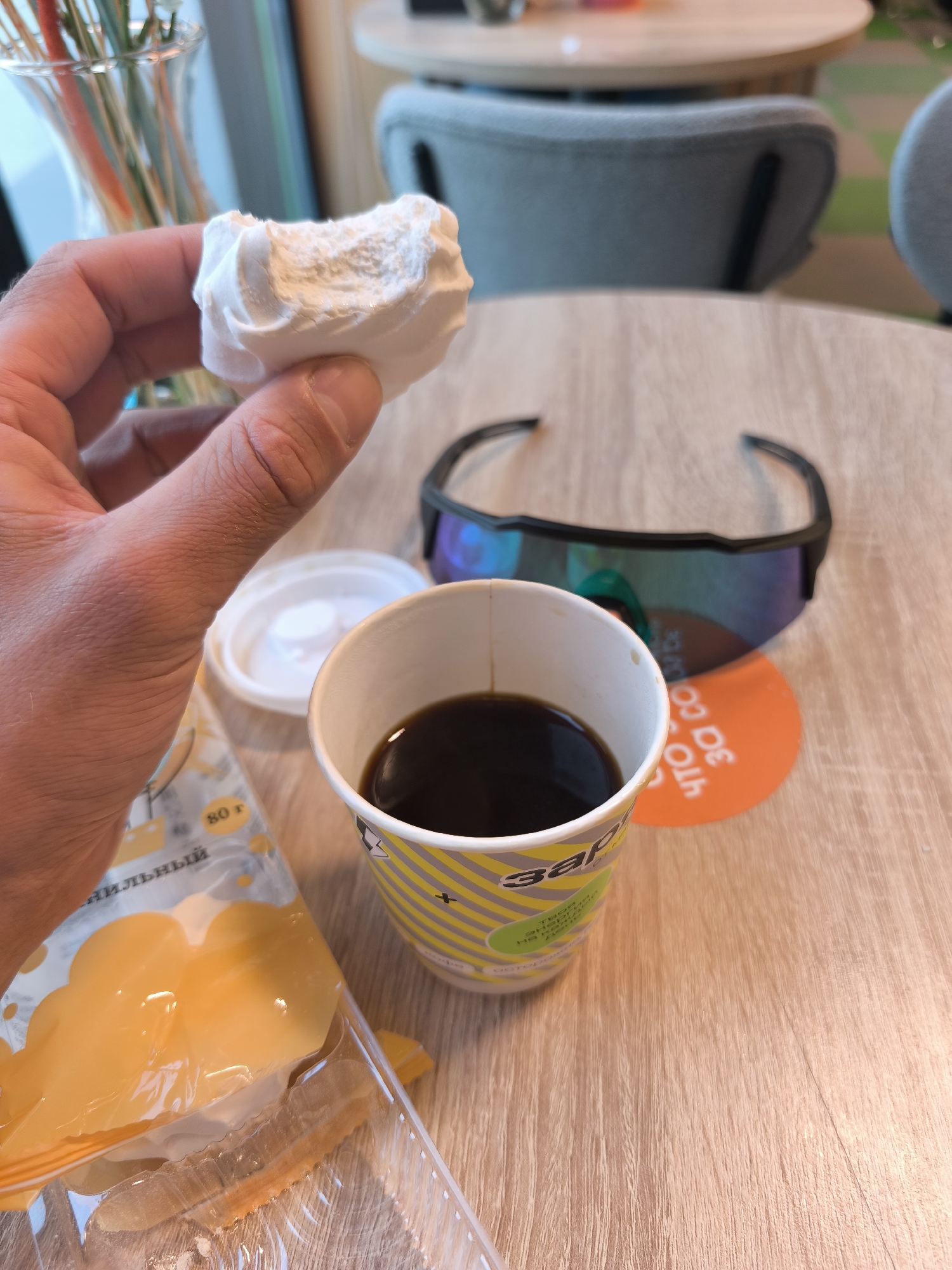 A close-up shot depicts a person holding a bitten piece of white marshmallow in their left hand. Adjacent to the hand is a plastic container filled with what appears to be orange marmalade candies. A disposable cup filled with a dark liquid, possibly coffee, is positioned near the marshmallow. A white lid for the cup rests beside it. Sunglasses with black frames are visible in the background, placed on a table. The table also features an orange coaster with Cyrillic text.
Based on the text on the cup, which appears to contain the word "Zaryadye," and the style of the disposable cup, the location is likely a café or eatery within or near Zaryadye Park in Moscow, Russia.