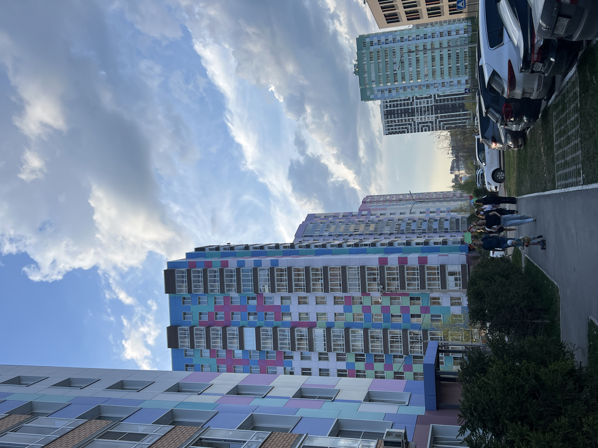 The image shows a row of tall, colorful apartment buildings in Perm, Russia, beneath a partially cloudy sky. The buildings are adorned with a patchwork of blue, purple, pink, and green panels, with rows of windows. A group of pedestrians is walking on a sidewalk alongside parked cars, with some greenery visible.