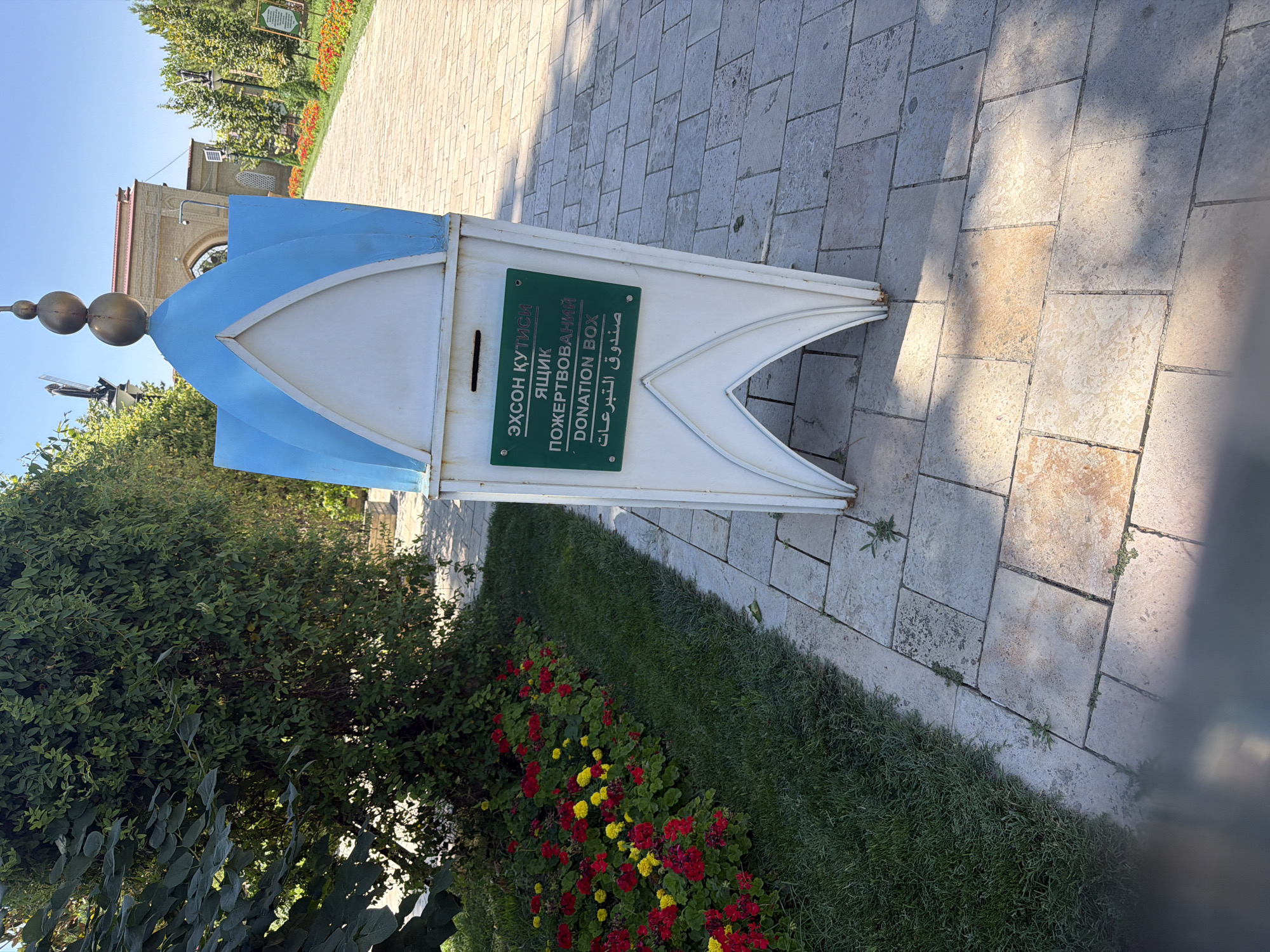 A donation box, shaped like a miniature blue and white mosque with a finial of three golden spheres, stands on a paved walkway next to a grassy area with red and yellow flowers. The donation box has a green sign with text in multiple languages, including "DONATION BOX". In the background, a building with a brick facade and an arched entrance is visible, along with trees and lampposts lining the walkway. The scene is set in a park or garden environment.