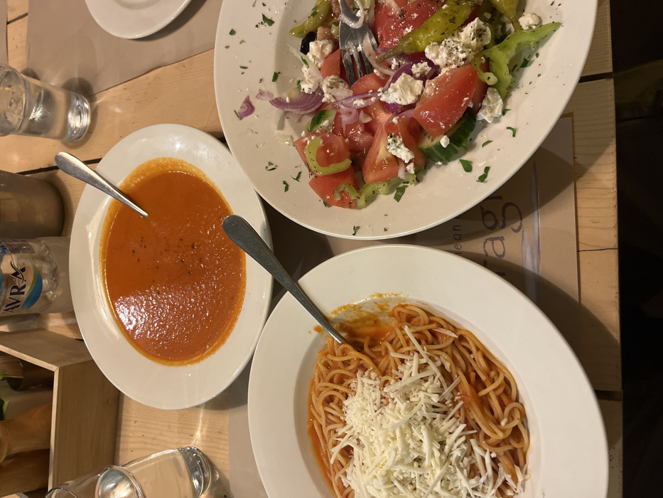 The image shows a table setting in a restaurant in Limenaria, Greece, at latitude 40.626 and longitude 24.577. The table is covered in a wooden surface with a light brown placemat. On the table are three white plates, two of which are filled with food. One plate contains a bowl of red tomato soup with a spoon beside it. The second plate holds a bowl of spaghetti with tomato sauce and a layer of grated white cheese. The third plate, to the right of the frame, contains a Greek salad with tomatoes, cucumbers, onions, feta cheese, and green peppers. To the right of the image is a partial view of a glass of water on the table. A fork is seen partially in the Greek salad. There is also a partially visible bottle of water in the upper left corner of the image.