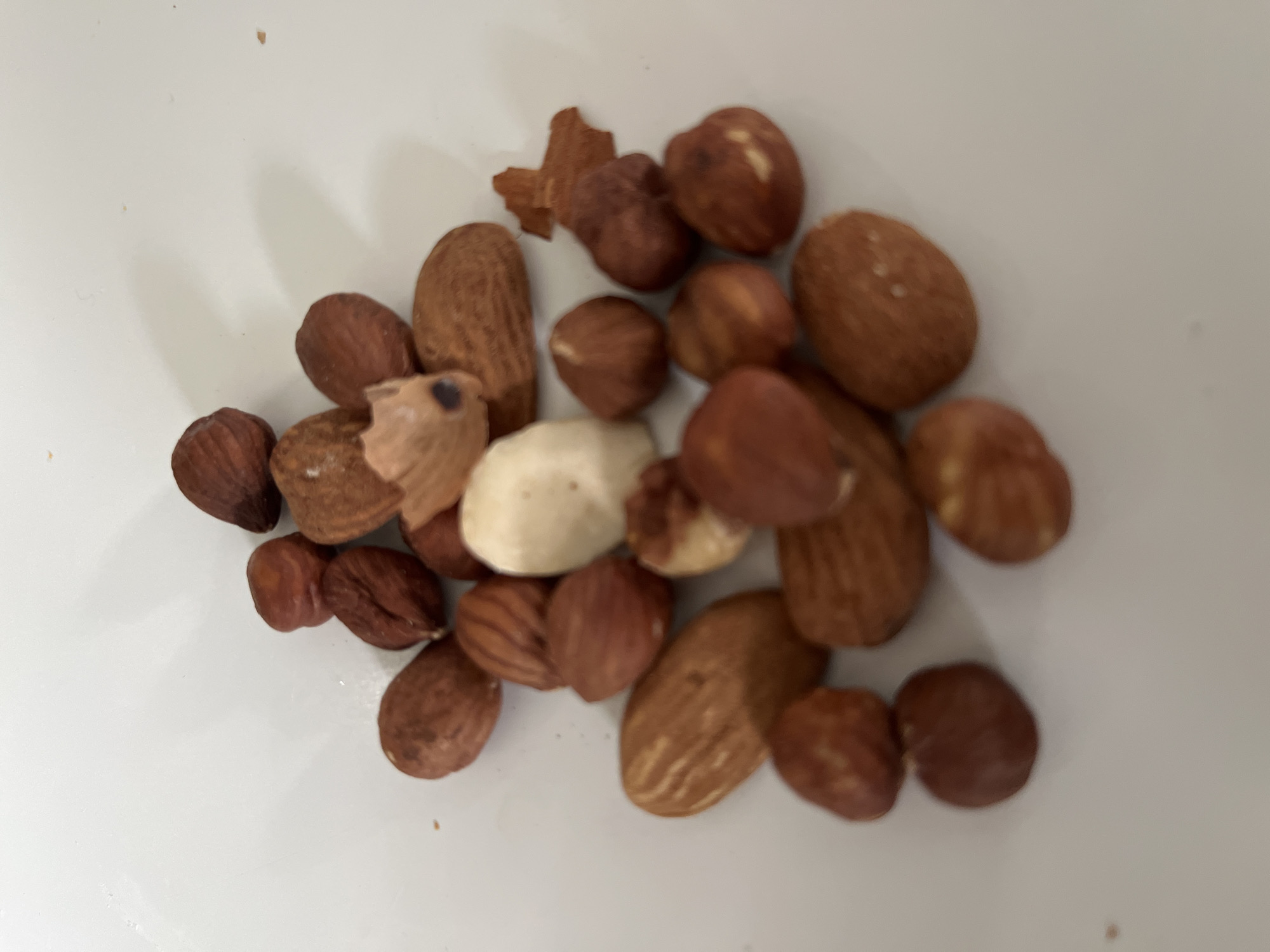 A pile of mixed nuts, including almonds and hazelnuts, is scattered on a white surface. One almond is notably pale, suggesting it may be blanched. The nuts are viewed from a top-down perspective. The scene does not provide any information about the location in Chambœuf, France.