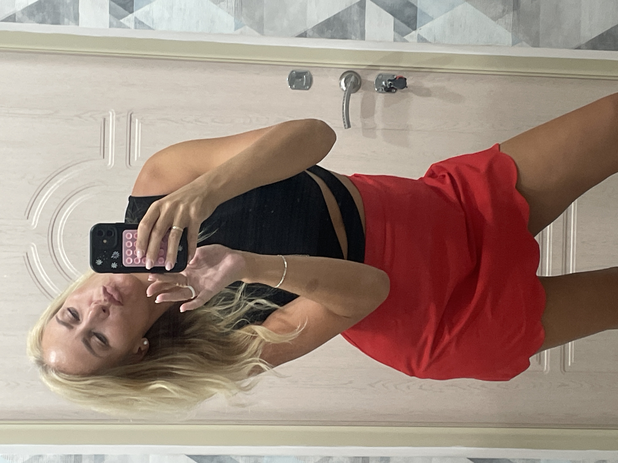 A woman with blonde hair is taking a mirror selfie. She is wearing a black crop top and a red skirt. The background appears to be a door with a handle and a keyhole. The woman is puckering her lips and holding her phone up to take the picture. The image was taken in Novorossiysk, Russia.