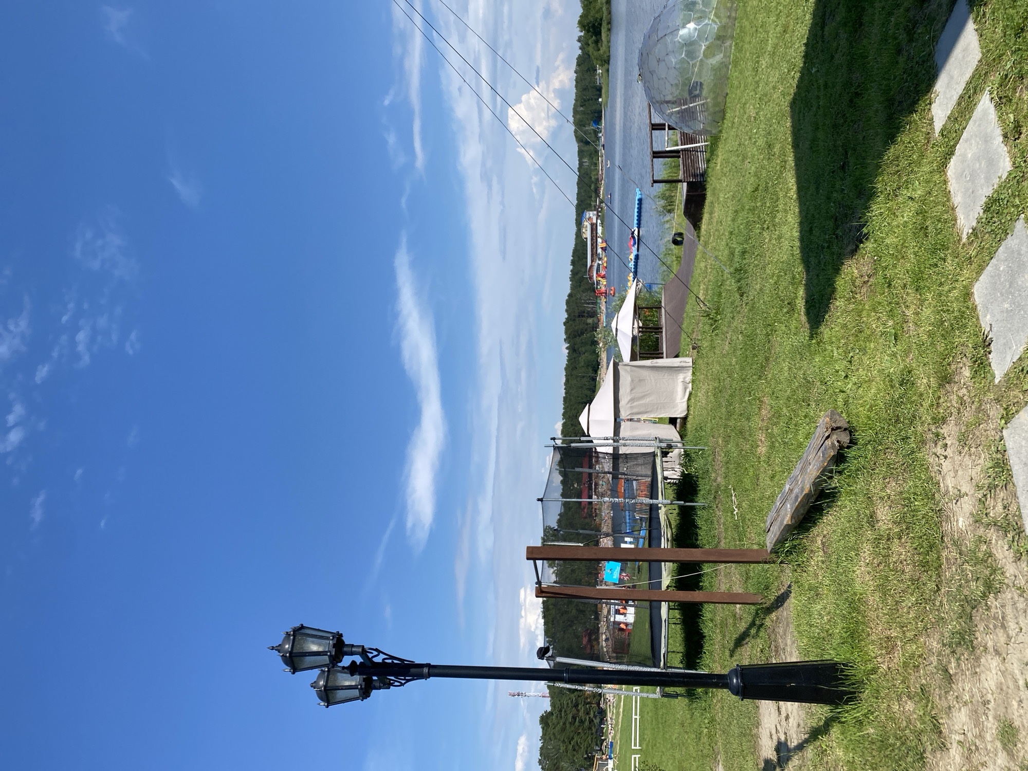 A low-angle, outdoor, full shot captures a sunny day at a recreational area by a lake. In the foreground, a black, ornate, double-headed street lamp stands to the left, its shadow cast across the green grass and patchy dirt path. To the right of the lamp, a rough-hewn log lies on the grass, and further back, a trampoline is set up with a safety net. Beyond the trampoline are several white tent structures and a transparent geodesic dome. The middle ground features a lake with a floating water park, various boats, and a wooden structure resembling a pier. In the background, a dense line of green trees borders the far side of the lake, beneath a bright blue sky dotted with white clouds. The scene depicts a leisure facility with various recreational activities available.