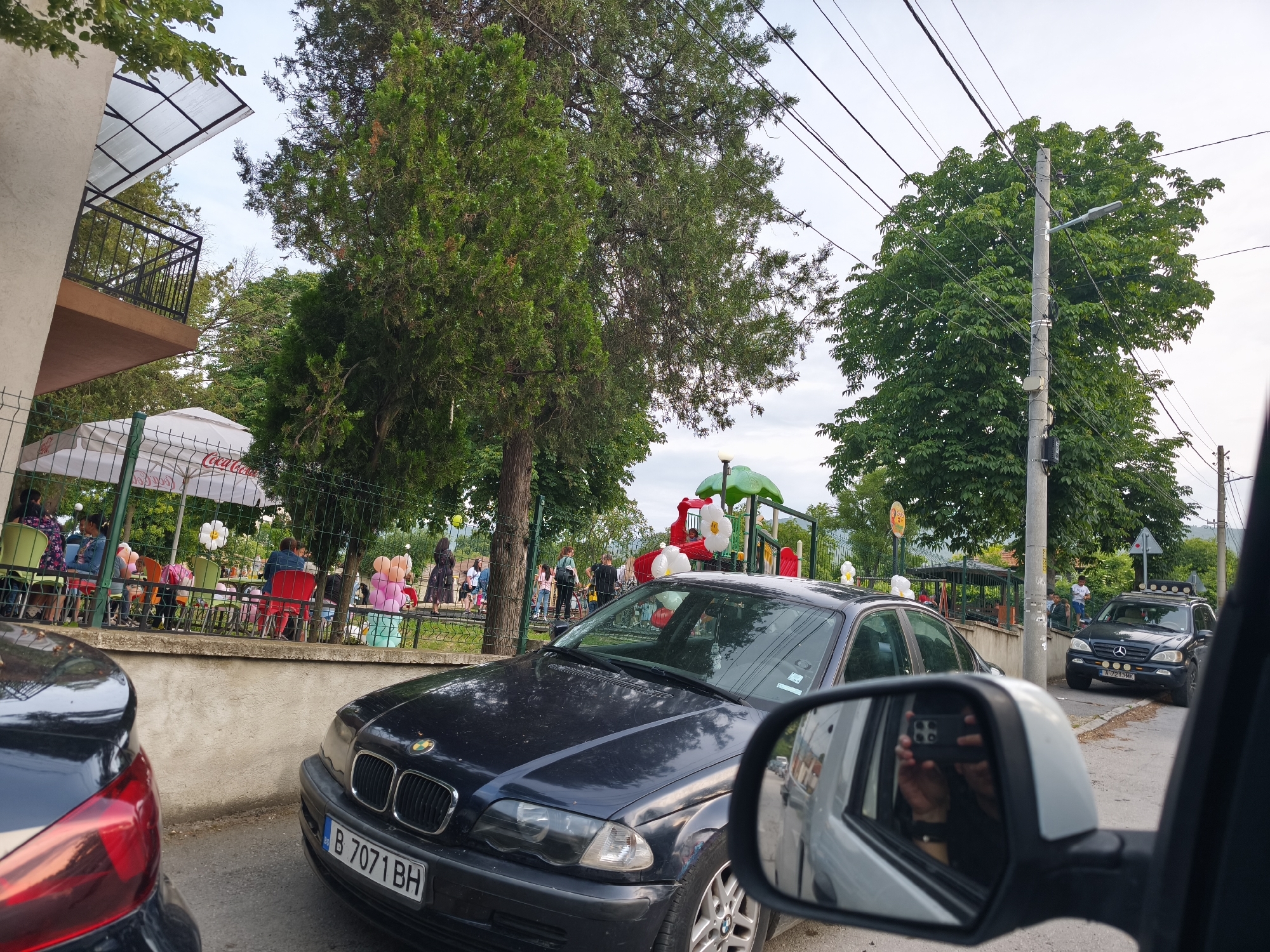 An outdoor scene captures a lively street or parking area adjacent to a cafe and a playground, suggesting a social gathering during the daytime. The sky is bright, possibly overcast.
In the immediate foreground, the rear of a dark vehicle with a prominent red taillight is visible on the left, and a car's side mirror on the right reflects a person holding a smartphone to take the picture. A dark blue BMW sedan (license plate B 7071 BH, indicating Bulgaria, Sofia region) is parked centrally, facing the viewer.
Beyond the cars, an outdoor cafe or restaurant seating area is visible, bustling with people seated at tables under a white umbrella bearing the "Coca-Cola" logo. A low wall and a metal fence separate this area from the foreground. Behind the cafe, a colorful playground with red, green, and yellow structures is adorned with white and pastel-colored balloons shaped like flowers or clouds, indicating a celebratory atmosphere, possibly a children's party. More people are gathered around the playground.
Abundant green trees frame the scene, and a utility pole with multiple power lines stands prominently on the right. Further down the street, a dark Mercedes-Benz M-Class SUV (license plate A 7213 MK, indicating Bulgaria, Blagoevgrad region) is parked. A building with a balcony and a translucent roof extension is partially visible on the far left. The overall impression is one of casual community activity.