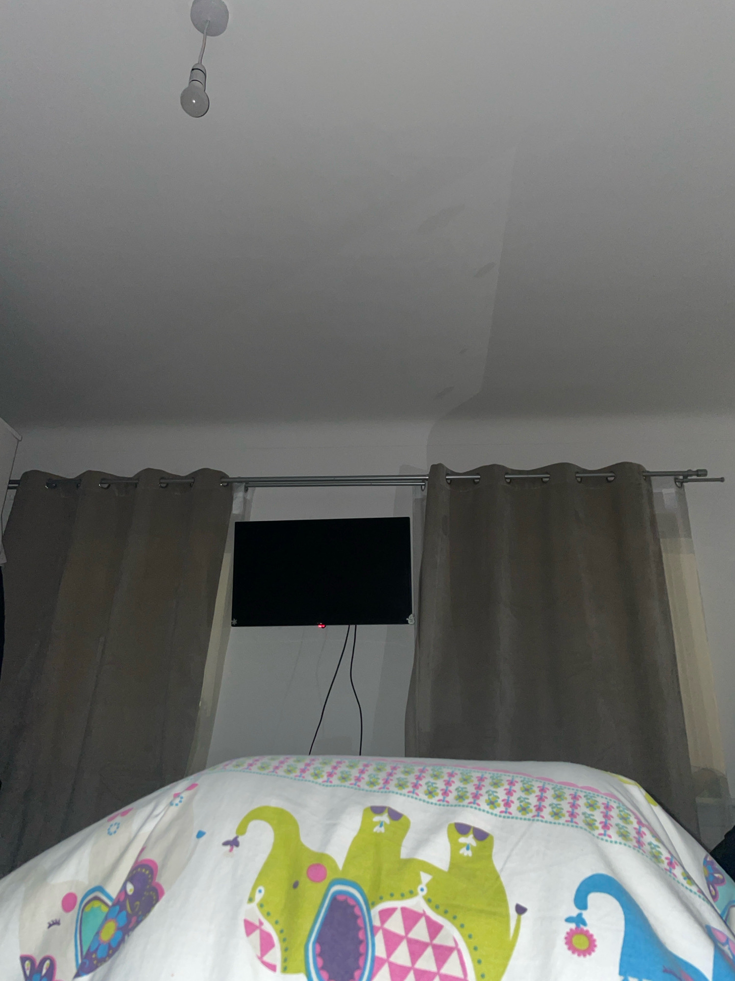 The image shows a bedroom interior, likely in Paisley, United Kingdom. In the foreground, a white duvet cover with a colorful pattern of stylized elephants and floral motifs is visible. Beyond the bed, a black flat-screen television is mounted on the wall, with a red indicator light visible. Two tan-colored curtains hang on either side of the television, obscuring the view of a window. The ceiling above has a modern light fixture with a visible bulb. The overall impression is that of a residential bedroom.