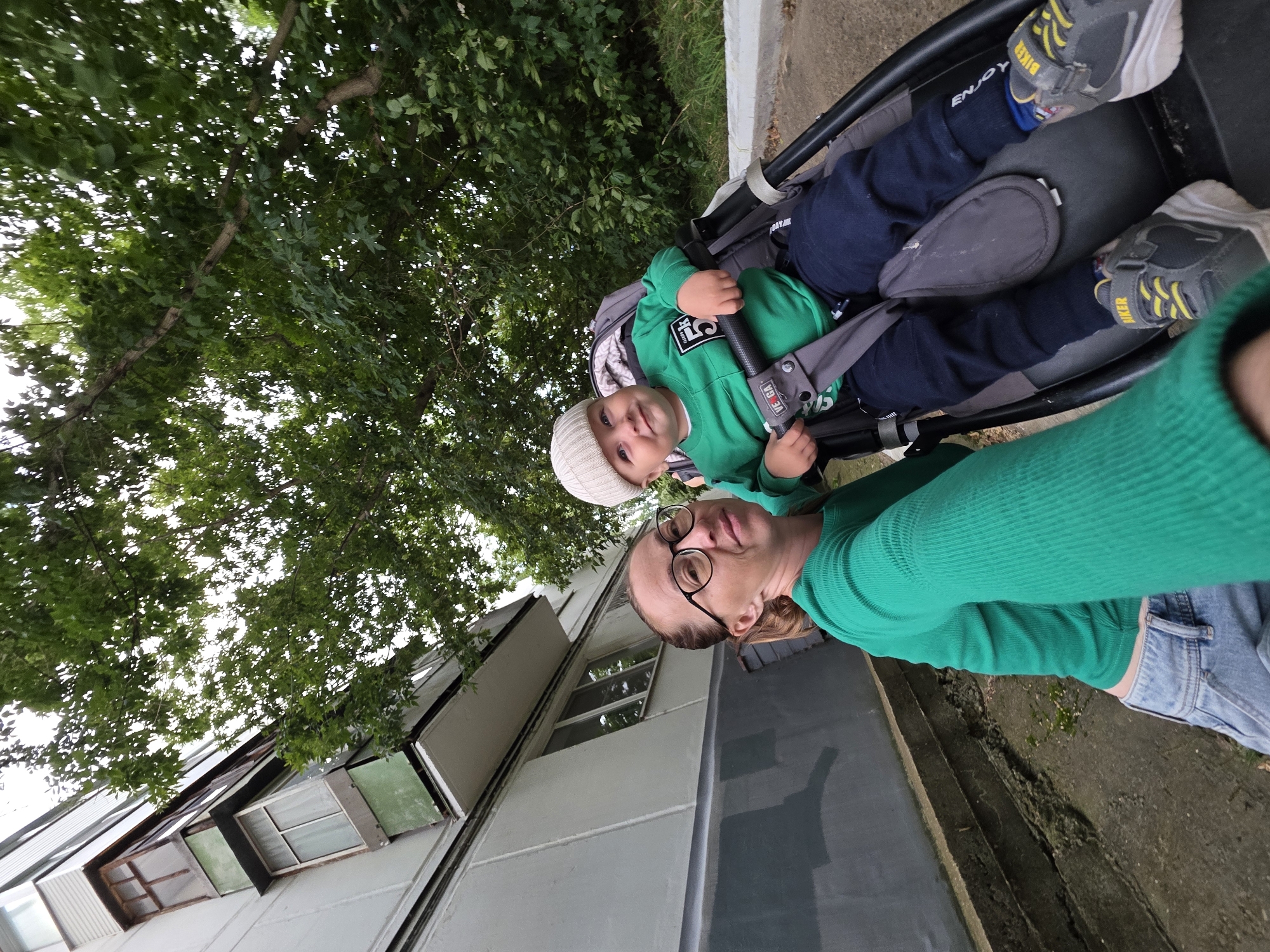 In Chaikovsky, Russia, a woman with glasses and a green top is taking a selfie. Beside her, in a stroller, is a young child wearing a white hat and a green sweatshirt. The background shows a building and trees.