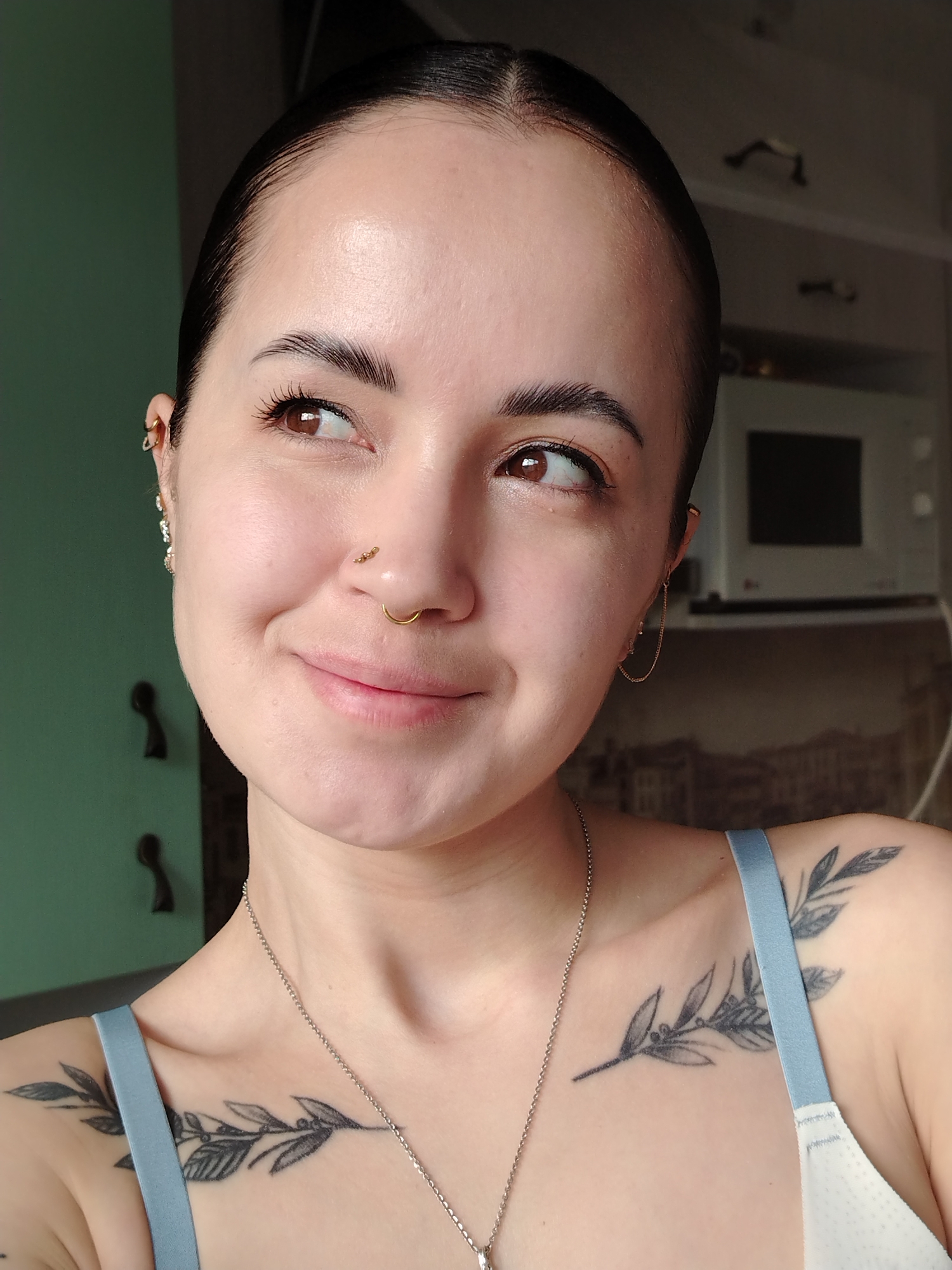 A brightly lit portrait features a young woman smiling subtly and looking towards her right. Her dark hair is pulled back tightly, revealing a clear complexion. She has full, dark eyebrows and brown eyes. Notable piercings include a gold hoop and a stud with three small gold beads on her left nostril, a gold cuff on her right ear, and a chain earring connecting two piercings on her left ear. A distinctive black ink tattoo of a leafy branch with berries is visible on her right collarbone and shoulder. She is wearing a light blue camisole or bra strap and a thin silver chain necklace.
The background suggests an indoor setting, possibly a kitchen or a room adjacent to one. A muted green/teal wall is visible to her left, while a lighter-colored wall with dark, curved handles is behind her to the right. A white microwave with a dark screen is partially visible in the upper right corner of the frame. The natural, bright lighting indicates it is daytime. Her expression conveys a sense of gentle contentment or thoughtfulness. No visible text is present in the image. The location context for the image is Noviy, Russia.