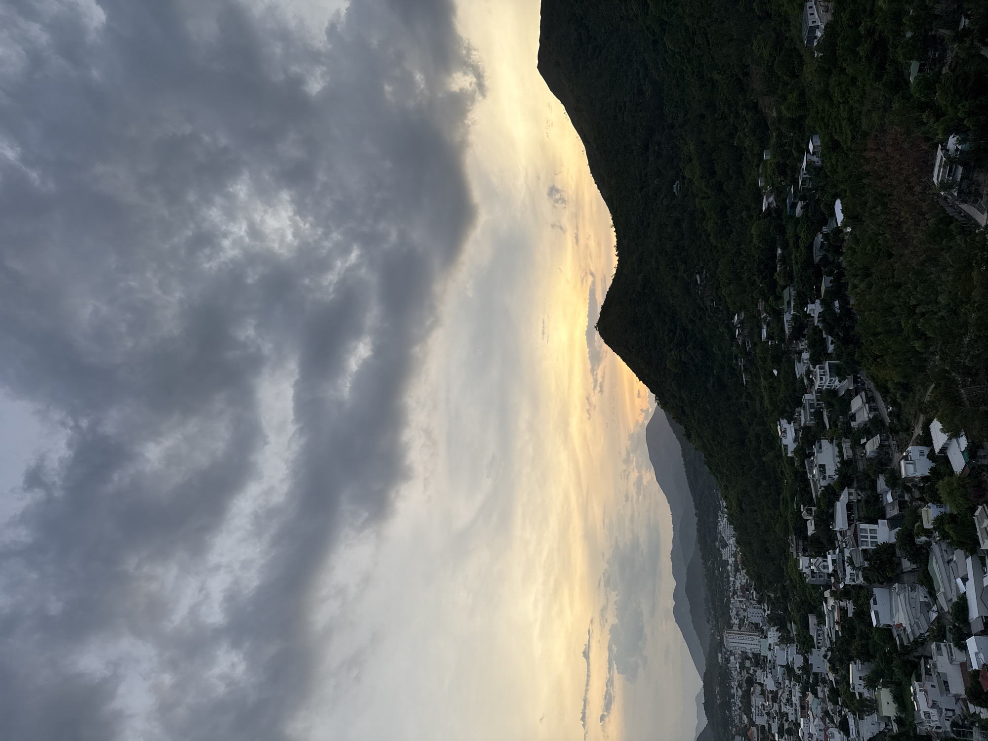 This is an aerial, wide-angle photograph of a coastal city and its surrounding mountainous landscape, likely taken during sunset in the Bac Natural Heritage Area, Trang, Vietnam. The sky is a dramatic blend of dark gray storm clouds dominating the upper half of the frame, with a soft, warm yellow and orange gradient near the horizon, suggesting the setting sun behind the clouds.
The foreground is dominated by a densely populated urban area, characterized by numerous white and light-colored buildings, varying in size and architectural style, interspersed with dense green vegetation. This urban sprawl extends across rolling hills. The buildings appear to be mostly residential, with some taller structures resembling apartment blocks or hotels visible in the distance. Roads are visible winding through the settlement.
Behind the city, lush green mountains rise sharply. The slopes are densely covered in trees, with the dark foliage contrasting against the lighter sky. The contours of several mountain ranges are visible, fading into a hazy purple in the far distance. One prominent, dark green mountain peak juts into the foreground from the right side of the frame. A few small structures are visible nestled within the forested slopes of these mountains. The overall impression is one of a city nestled between the sea (implied by the coastal location but not visible) and rugged, verdant terrain, under a moody sky. There are no people or animals discernible in the image.