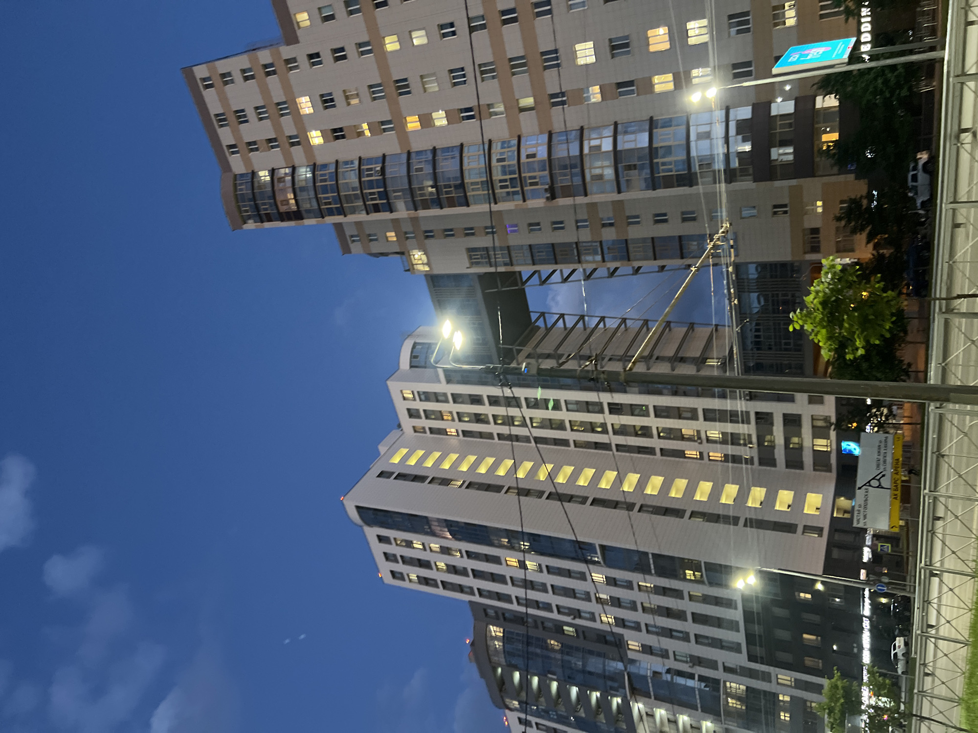 The image captures a view of modern high-rise apartment buildings in Dachnoe, Russia, during twilight. The sky is a deep blue with scattered clouds. The buildings are tall and feature a mix of light-colored facades and extensive glass windows, many of which are illuminated from within. A prominent architectural feature is a connecting bridge-like structure between two of the buildings. In the foreground, a street with cars is visible, separated from the viewpoint by a metal fence and a grassy area. Streetlights and power lines are also present in the scene. Signs in Russian are visible on the side of the road. The overall impression is of a residential urban environment at dusk.