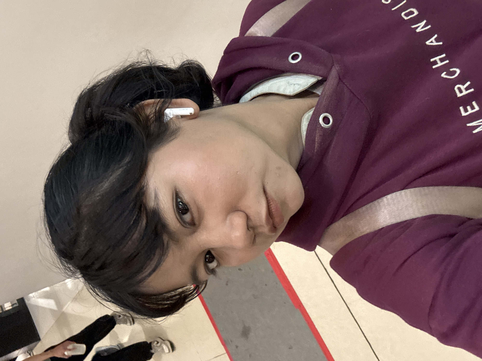 A young woman with dark hair pulled back into a ponytail is taking a selfie indoors, likely in a public space. She is wearing a maroon hoodie with a light-colored collar and a light-colored strap crossing her shoulder. She has a white earbud in her right ear. In the background, two other individuals are partially visible, one holding a white cup and the other wearing sneakers. The floor appears to be tiled with a gray mat outlined in red. Based on the context of similar images, the location is likely a mall in North Jakarta, Indonesia.