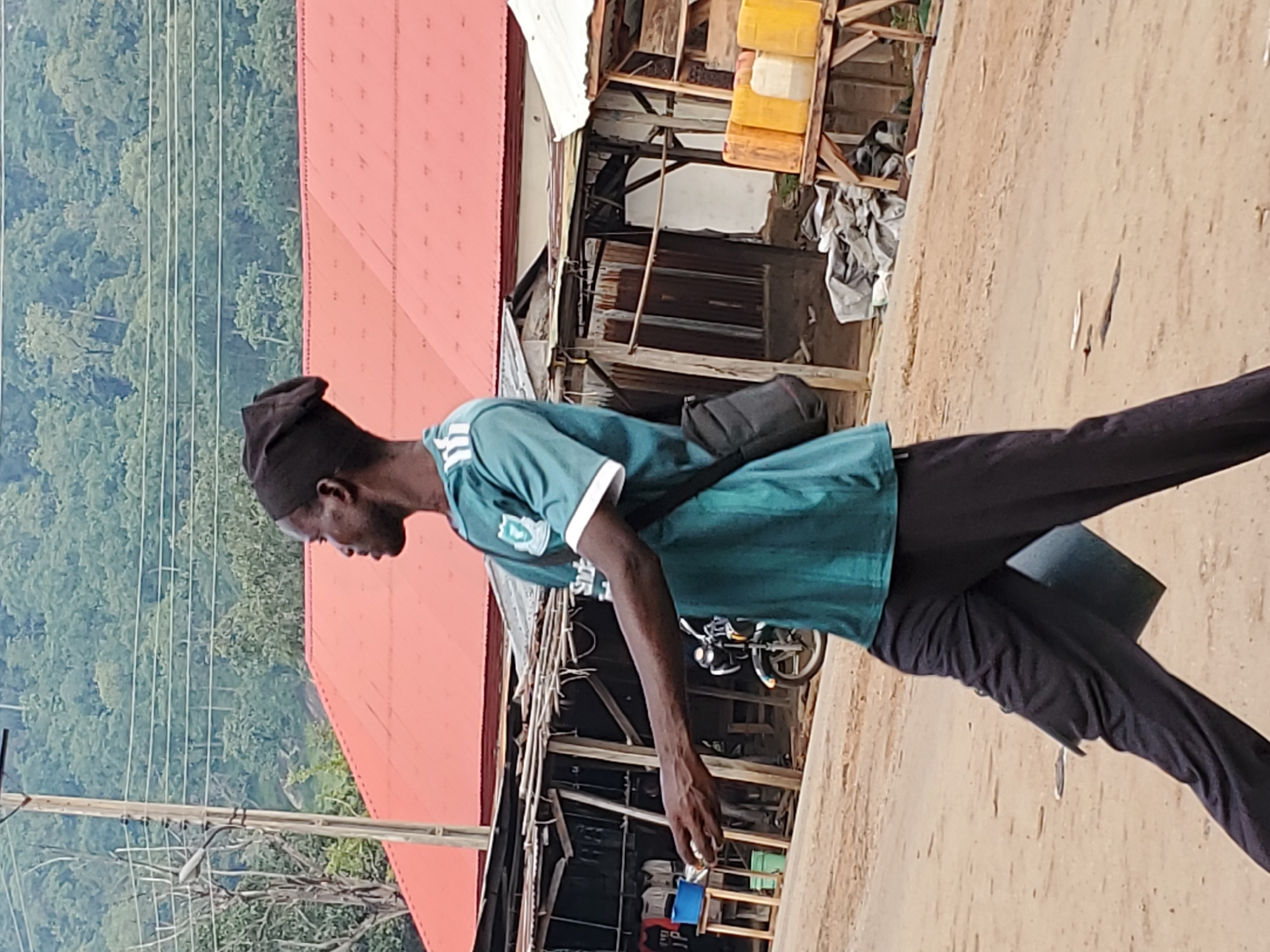 In Mararaba, Nigeria, a man walks along a dirt road next to a building with a red roof and partially constructed structures. The man wears a dark cap, a green and white shirt, and dark pants. He carries a dark bag over his shoulder and holds a small object in his hand. Behind the buildings are trees and power lines.