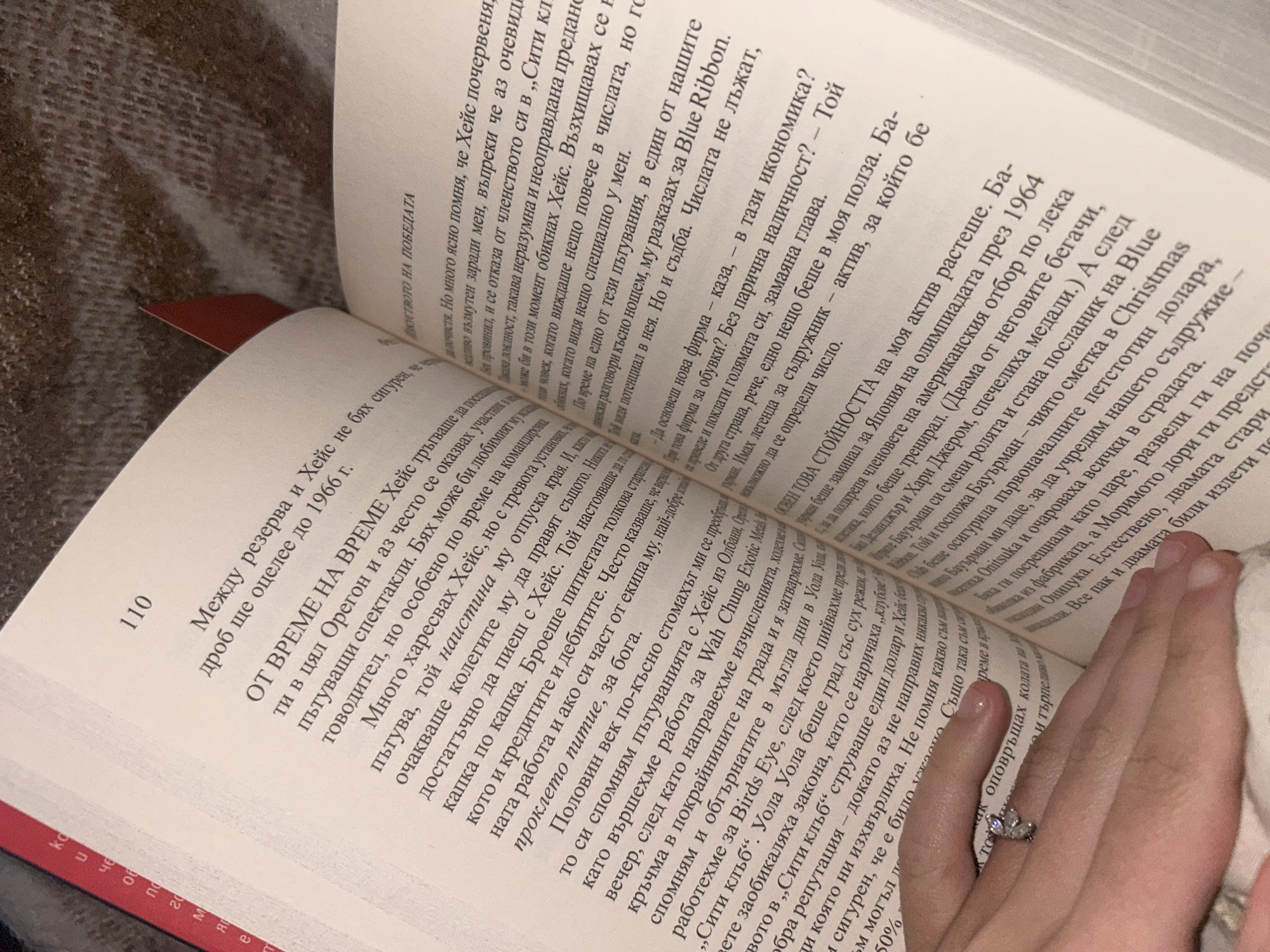 A close-up shot features a book opened, exposing two pages of Bulgarian text. A hand, adorned with a ring, rests on the right page. The book is lying on a soft, textured surface. The scene is well-lit, providing clear visibility of the text and the hand.