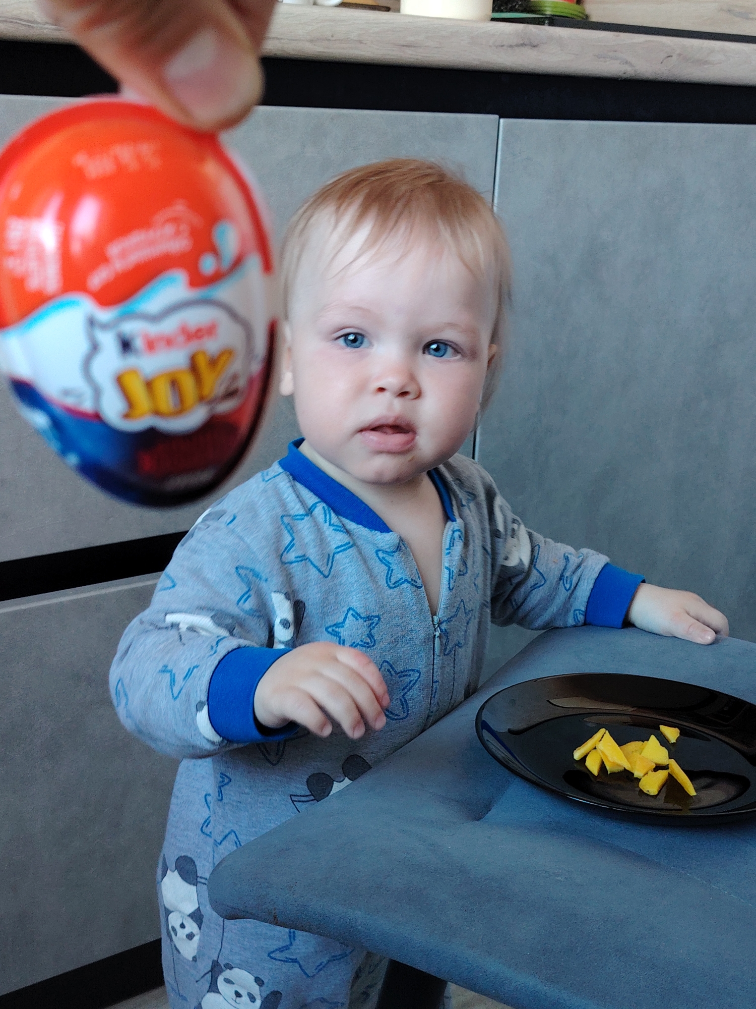 An indoor scene in Chaikovsky, Russia, features a baby and an adult's hand presenting a Kinder Joy egg. The baby, with light blonde hair and bright blue eyes, is positioned at a table or high chair, looking directly forward with a curious expression and slightly open mouth. The baby is wearing a gray full-body romper with blue accents, patterned with stars and pandas. Their left hand rests on the dark blue upholstered surface of a chair or high chair.
In the foreground, slightly out of focus, an adult's hand holds up a Kinder Joy egg, predominantly orange and white, with the brand name "Kinder Joy" clearly visible. On the dark blue surface in front of the baby, a black plate contains several small, irregularly shaped yellow pieces of food, possibly fruit or cheese. The background shows gray kitchen cabinets or wall panels. The lighting suggests an indoor setting during the daytime. The overall impression is a moment of interaction between the adult and the baby, with the Kinder Joy egg as a focal point.