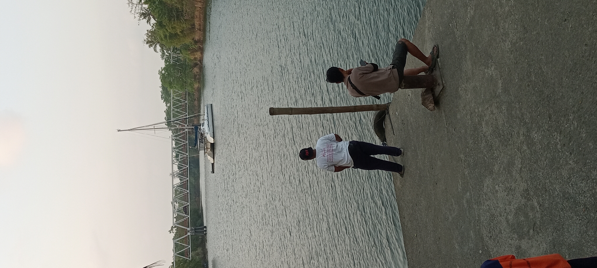 A wide-angle, vertical, eye-level shot captures a tranquil river scene in Kalipucang, Indonesia, with two men present on a concrete pier in the foreground. The sky is a pale, overcast grey, indicating early morning or late afternoon. A prominent, white catamaran sailboat with two masts is anchored in the river, to the left of a modern, silver truss bridge that spans the water. Lush green trees line the riverbanks. The concrete pier where the men are standing is rough and textured, with a single wooden post rising vertically in the middle of the scene.

The man on the left is standing, facing away from the camera, with his hands clasped behind his back. He is wearing a white t-shirt with text on the back and dark pants. The man on the right is seated on a dark, metal bollard, also facing away from the camera, engrossed in his mobile phone. He is wearing a brown t-shirt and dark shorts. The water is calm with subtle ripples, reflecting the overcast sky. To the far left, a small portion of another person wearing an orange and blue shirt is visible, suggesting their presence just outside the main frame.