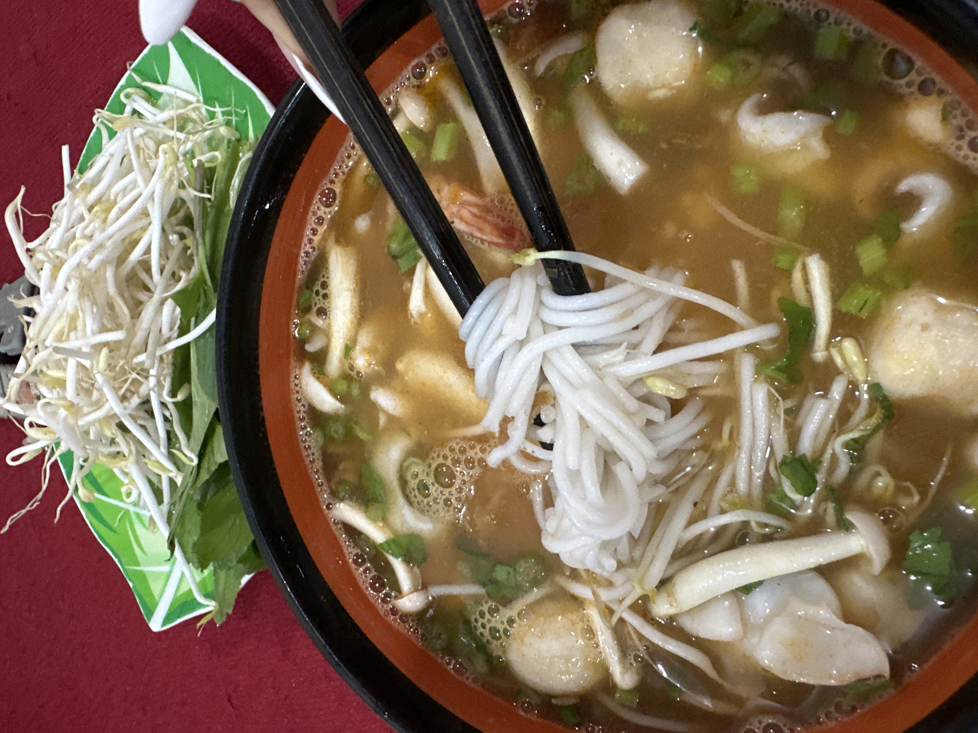 This close-up image shows a person enjoying a bowl of noodle soup, characteristic of Vietnamese cuisine. A hand is visible at the top, holding black chopsticks that are lifting a substantial amount of white rice vermicelli noodles from the bowl.
The soup is presented in a dark bowl with an orange rim and contains a rich, amber-colored broth dotted with small bubbles. Numerous ingredients are visible within the broth, including several round, light-colored items resembling fish balls, small white mushroom-like pieces, and chopped green onions. Other white, curved pieces, possibly seafood like squid, are also present, along with a hint of a reddish-orange shrimp or prawn. Bean sprouts are mixed into the soup, adding texture.
To the left of the main bowl, a small green and white plate holds additional fresh garnishes, primarily a generous pile of bean sprouts and some green leafy herbs, likely mint or basil, ready to be added to the soup as desired. The entire meal rests on a vibrant red surface, possibly a tablecloth or table. The scene captures a casual dining experience, likely indoors. While no specific location cues are visible in the image itself, the style of the meal is consistent with food found in places like Mũi Né, Vietnam, where such noodle soups are a staple. No text is visible, and the time of day or emotions cannot be determined from the close-up shot.