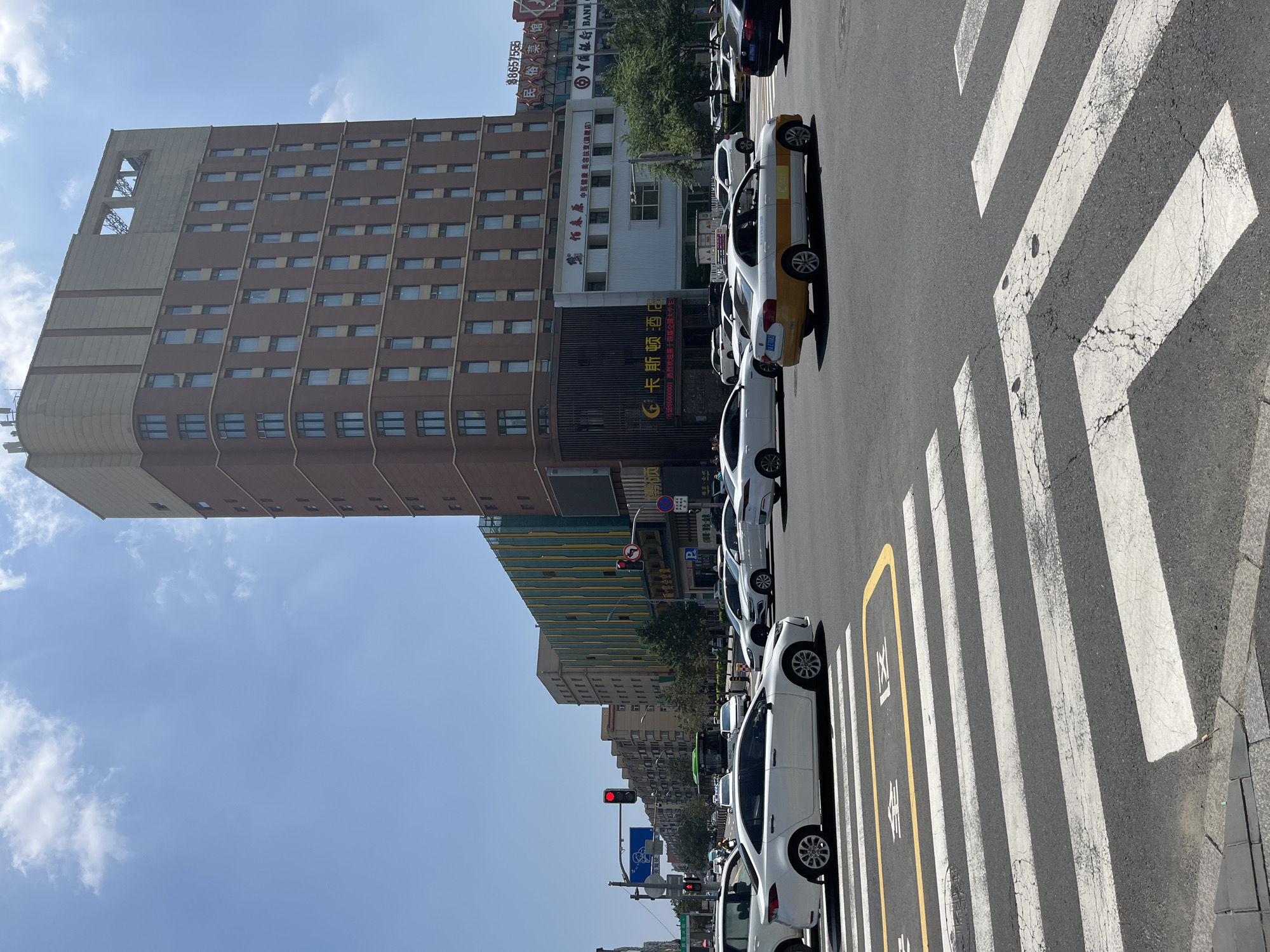The image captures a street scene in Changchun City, China, under a clear blue sky with scattered clouds. The foreground features a pedestrian crossing with white stripes on asphalt. In the middle ground, several cars are navigating the intersection, including a white sedan, a yellow and white taxi, and a black sedan. Beyond the intersection, a tall, multi-story building with a reddish-brown brick facade and cream-colored trim dominates the skyline. This building has numerous windows and appears to be a hotel or office building. To the left of this structure, a colorful building with a green and yellow striped facade is visible, along with other smaller buildings and trees. Traffic lights are visible, indicating an active urban environment. The overall impression is that of a bustling city intersection on a sunny day.