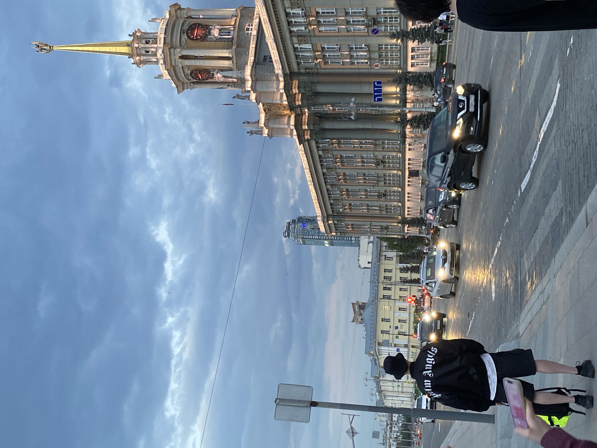 The image depicts a street scene in Yekaterinburg, Russia, featuring the city administration building with its clock tower on the left, and cars lining the street on the right. The sky above is filled with clouds. In the foreground, two people are visible, one holding a mobile phone. The image is rotated counter-clockwise.