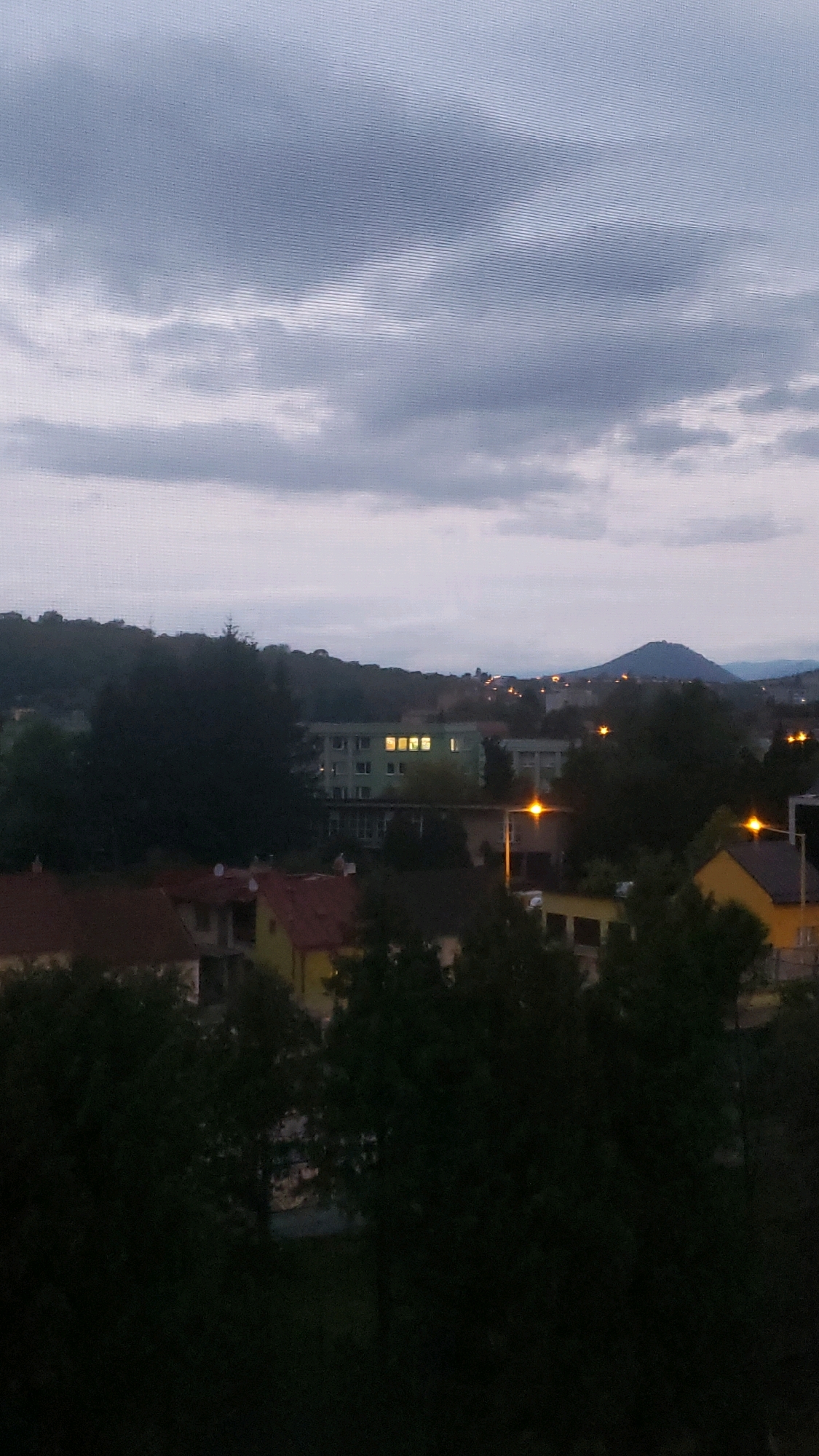 The image depicts a twilight scene over a residential area in Prešov, Slovakia. The sky is filled with overcast clouds, casting a muted light over the landscape. In the foreground, dark silhouettes of trees dominate, obscuring the lower parts of the buildings.
Further back, several houses with red and yellow roofs are visible, interspersed with more trees. A multi-story building with illuminated windows stands out among them. The background reveals a rolling landscape with more buildings and streetlights, suggesting the edge of the city. A prominent hill or small mountain is visible in the distance, with scattered lights indicating development on its slopes. The overall impression is one of a quiet evening in a suburban or semi-rural part of Prešov.
