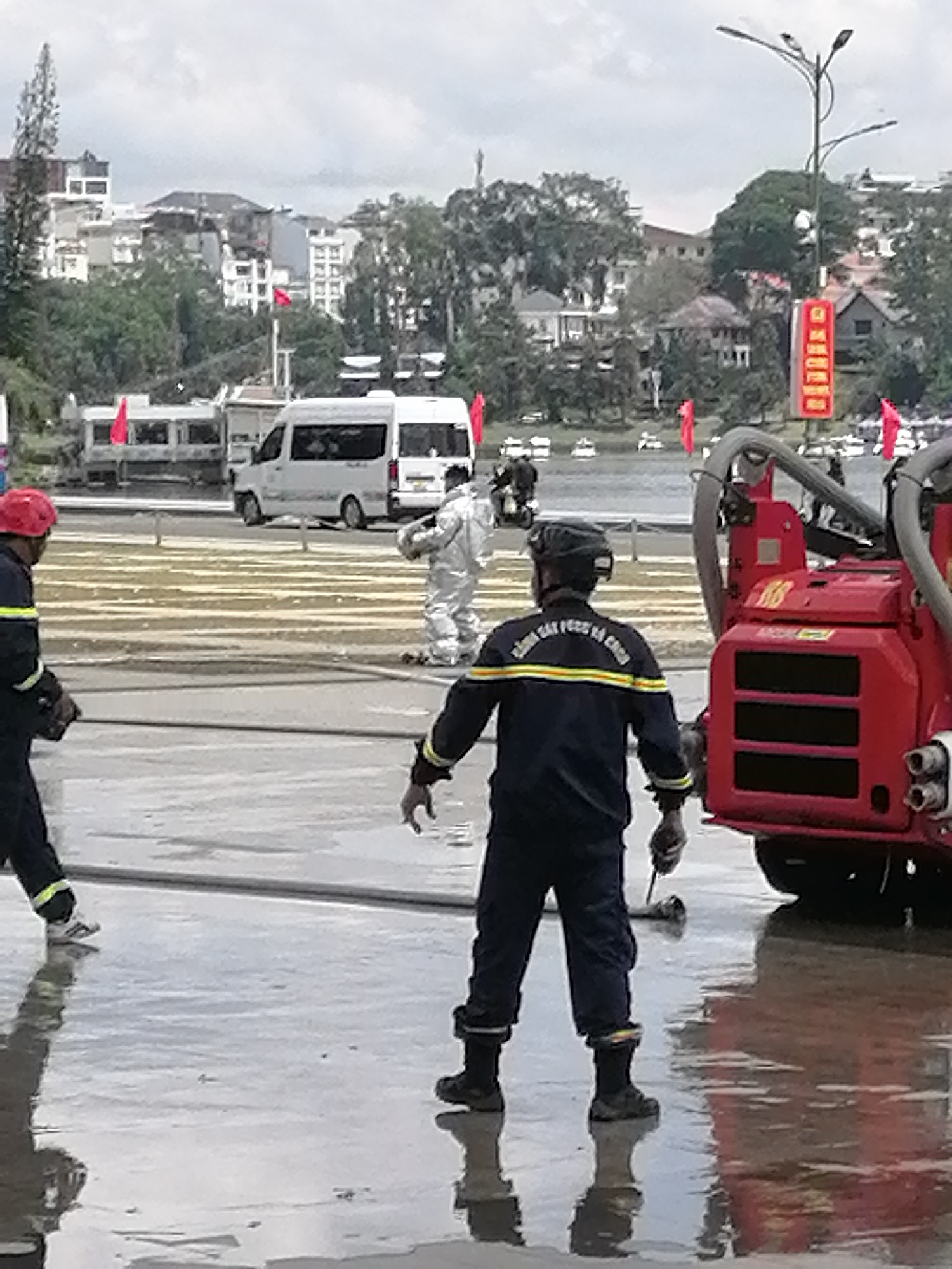 The image depicts an outdoor scene, likely on a street or open area, with several individuals present, possibly related to a public event or emergency response. In the foreground, two individuals in dark uniforms with reflective yellow and blue stripes are visible. One is facing away from the camera, wearing a helmet and holding what appears to be a tool. The other is partially visible on the left, also wearing a helmet. Behind them, a red fire apparatus with various hoses and components is parked on a wet, reflective surface, indicating recent rainfall or water presence. Further in the background, another person in a white protective suit is standing. In the mid-ground, a white minivan and a bus are parked. The background reveals a cityscape with buildings and trees on a hillside, consistent with the terrain of Da Lat, Vietnam. Red flags are visible throughout the scene, suggesting a local event or civic display. The overall atmosphere is somewhat overcast, with a muted color palette.